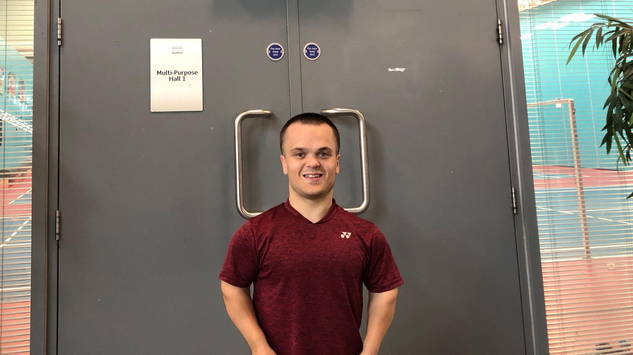 Jack Shephard smiles at the camera. He wears a red t-shirt and stands in front of a double door with a sign that reads 'multi purpose hall 1'.