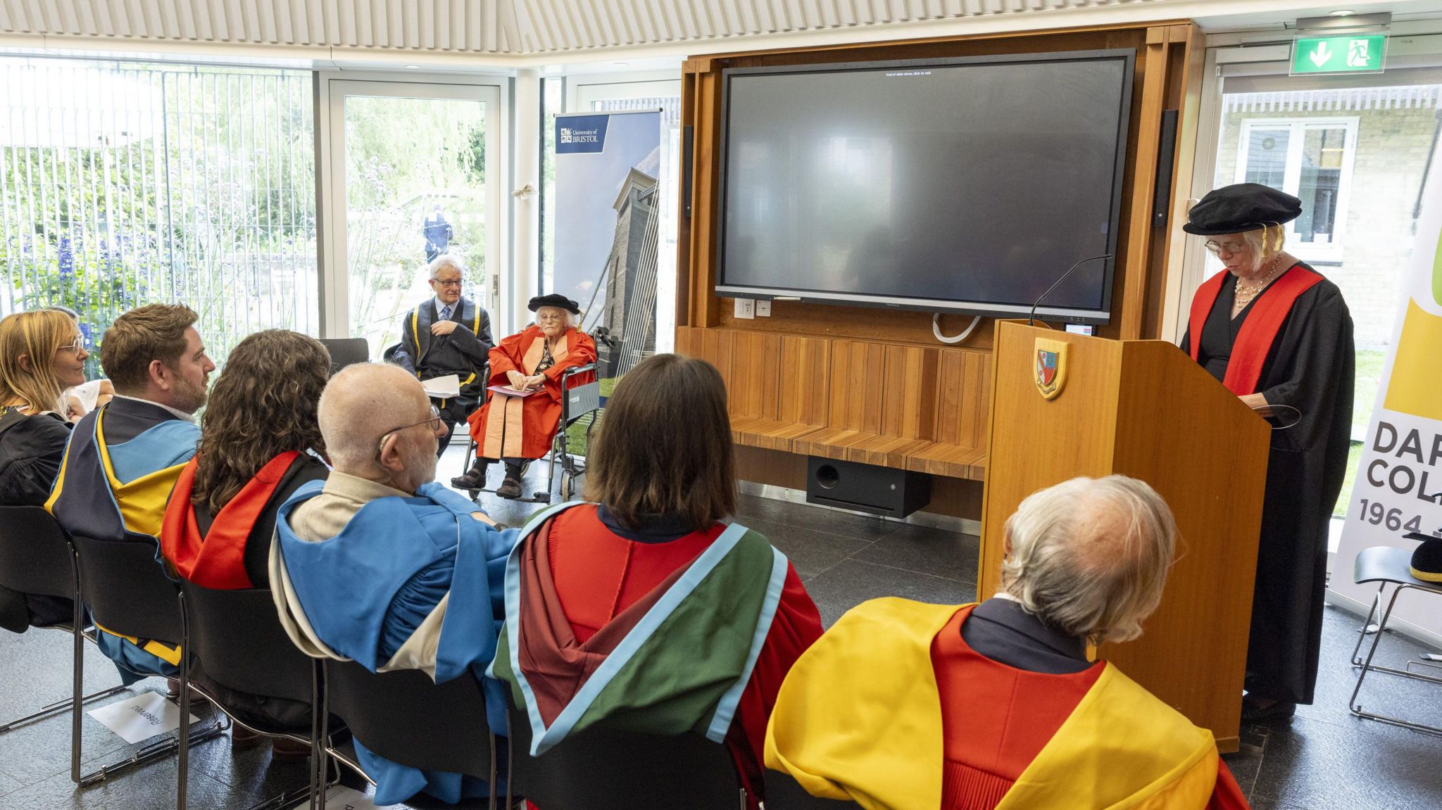 A row of people dressed in colourful graduation gowns sit in front of a lectern and a big screen