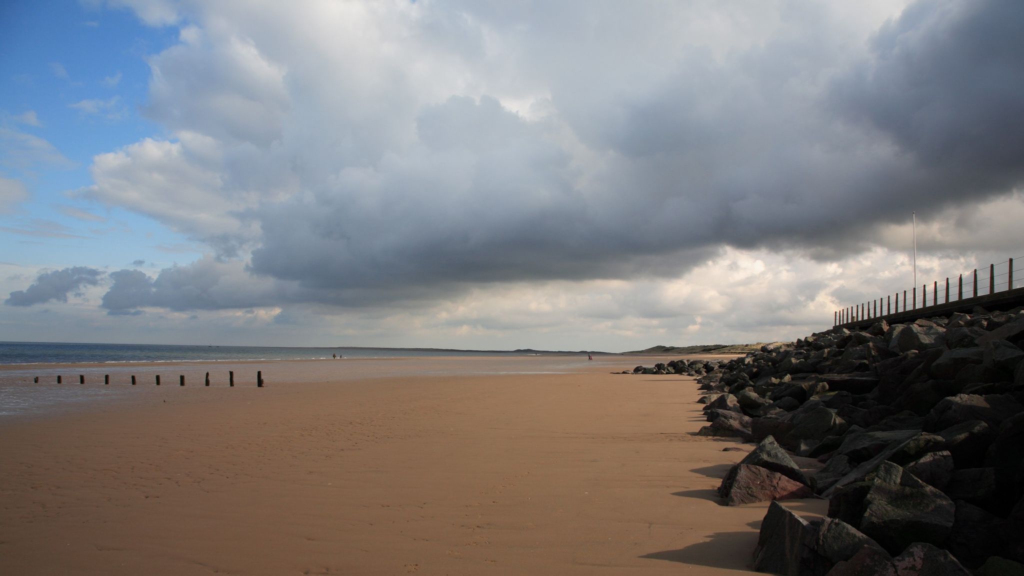 A stretch of empty beach with dark rocks piled up to the right against a sea barrier, and the sea to the left. 