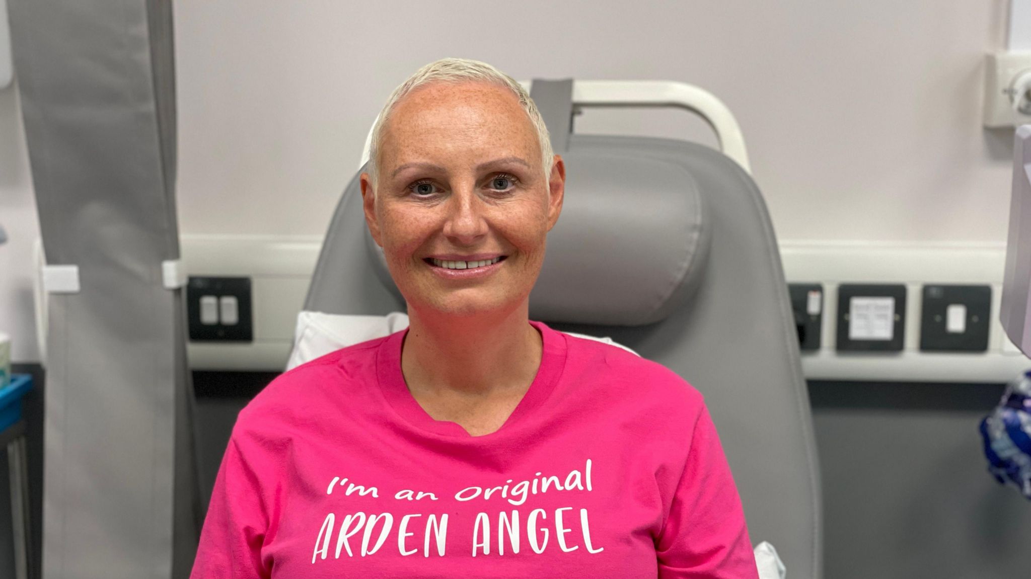 Sam Pooler sitting in a chemotherapy chair. She wears a pink T-shirt with "I'm an original Arden Angel" written on it. The chair she sits in has grey padding and is in front of a grey wall with several power sockets visible in the wall