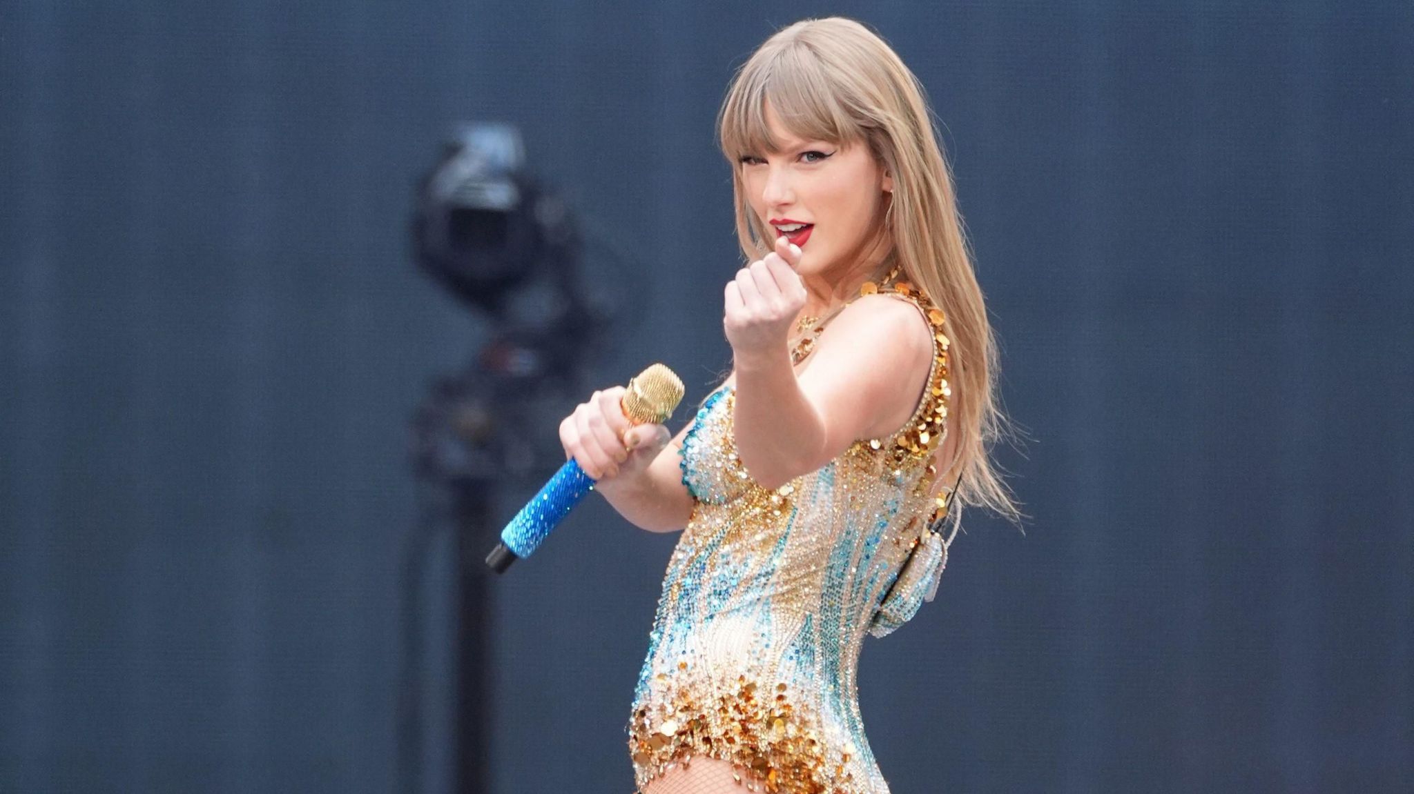 Taylor Swift performing in a sparkling gold and blue leotard, holding a blue and gold microphone and looking at the camera