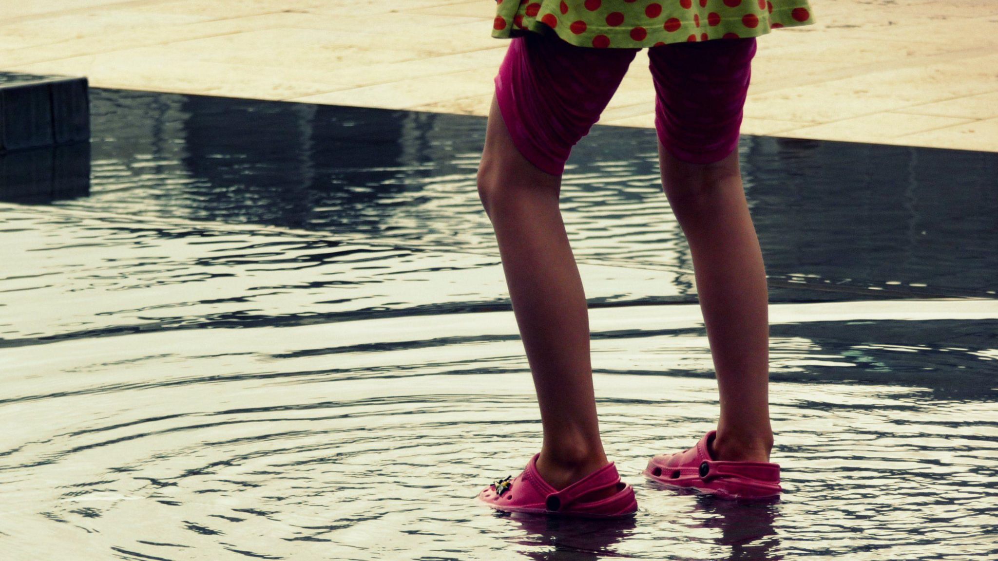 A child wearing a green and pink polka dot dress, pink shorts and bright pink crocs, pictured splashing the fountain at Millennium Square. 