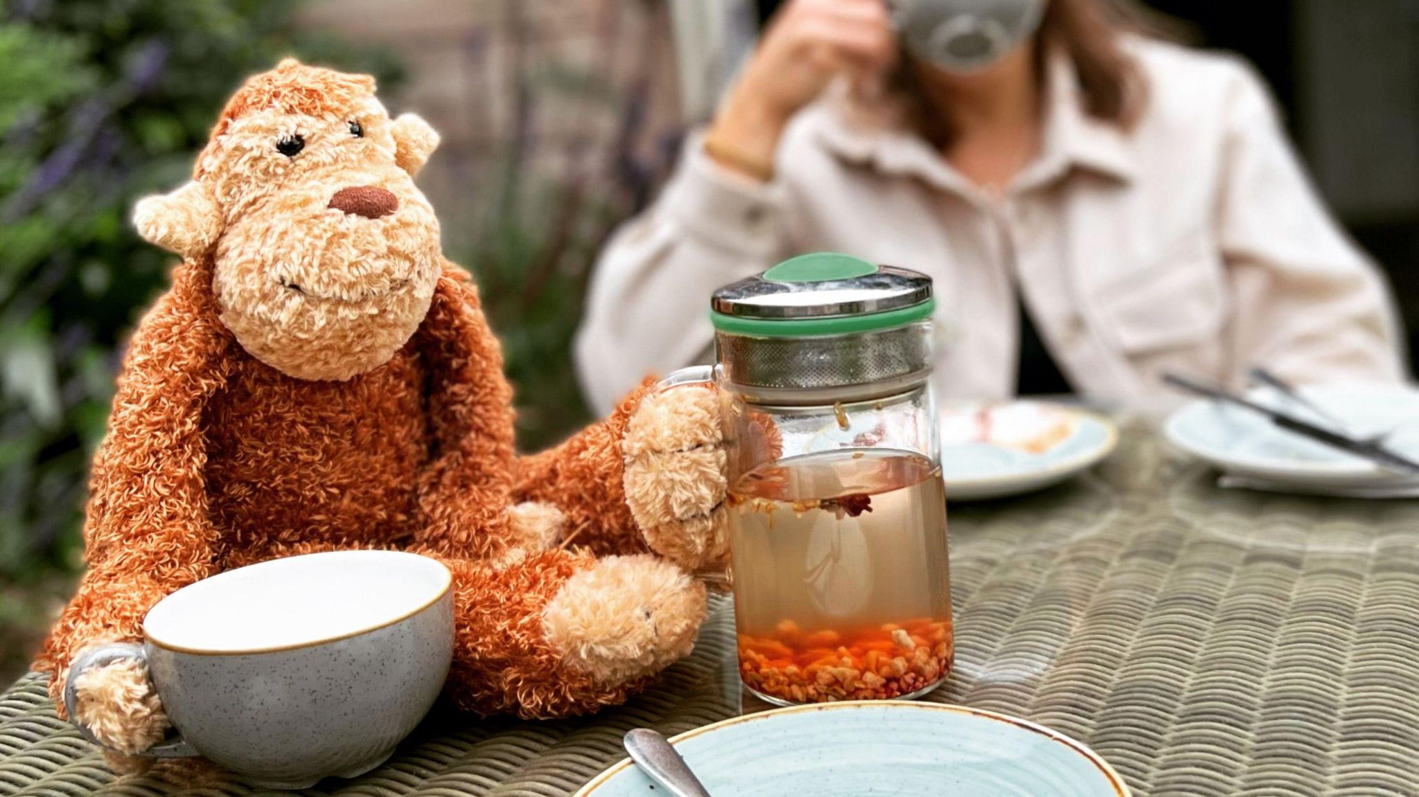 A toy monkey sits on a table with its paw touching a cup, as if it was to drink from it. 