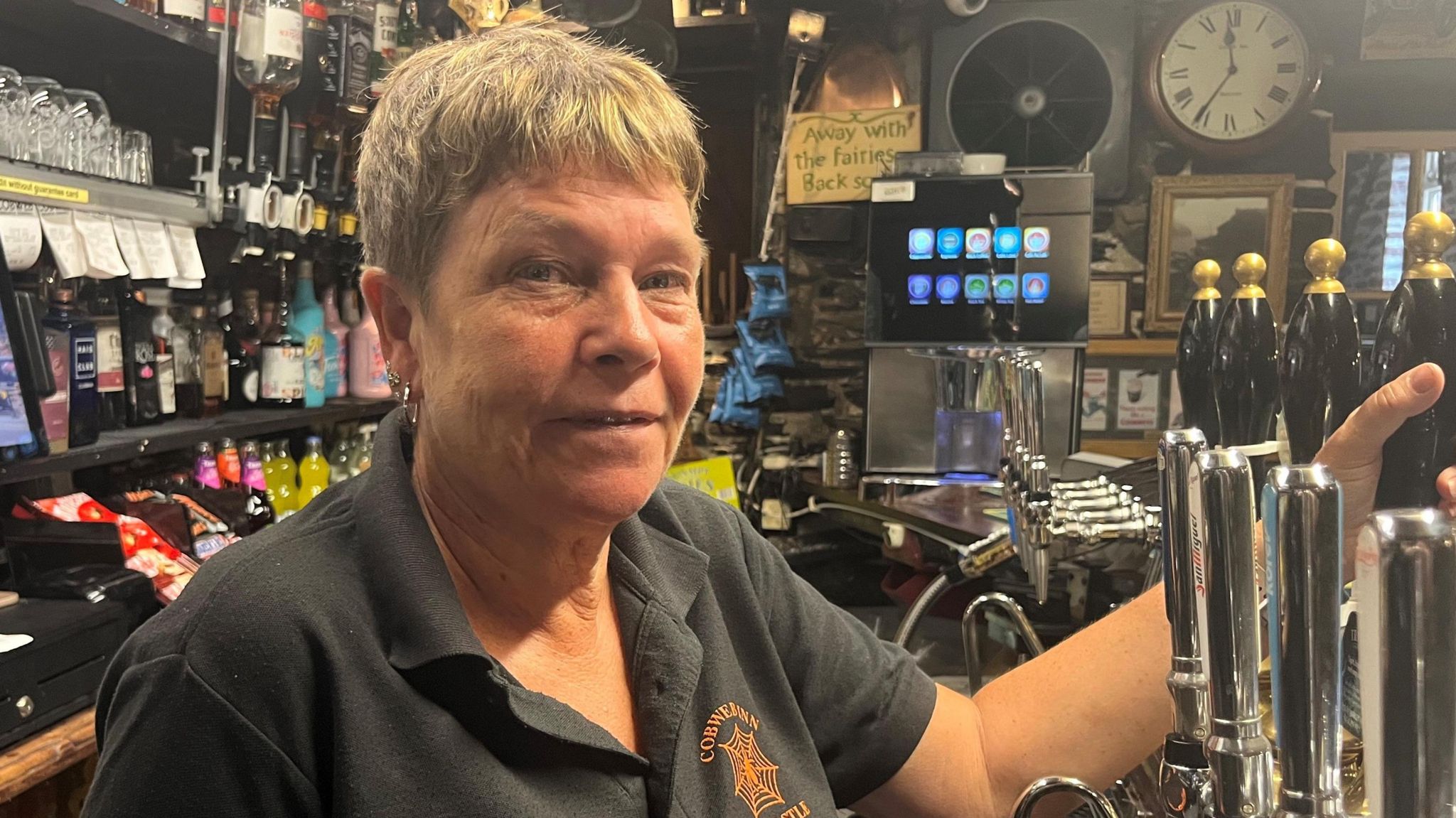 Cobweb Inn landlady Tracy Bright is standing behind the bar wearing a black polo shirt with the pub's motif on. She has short hair.