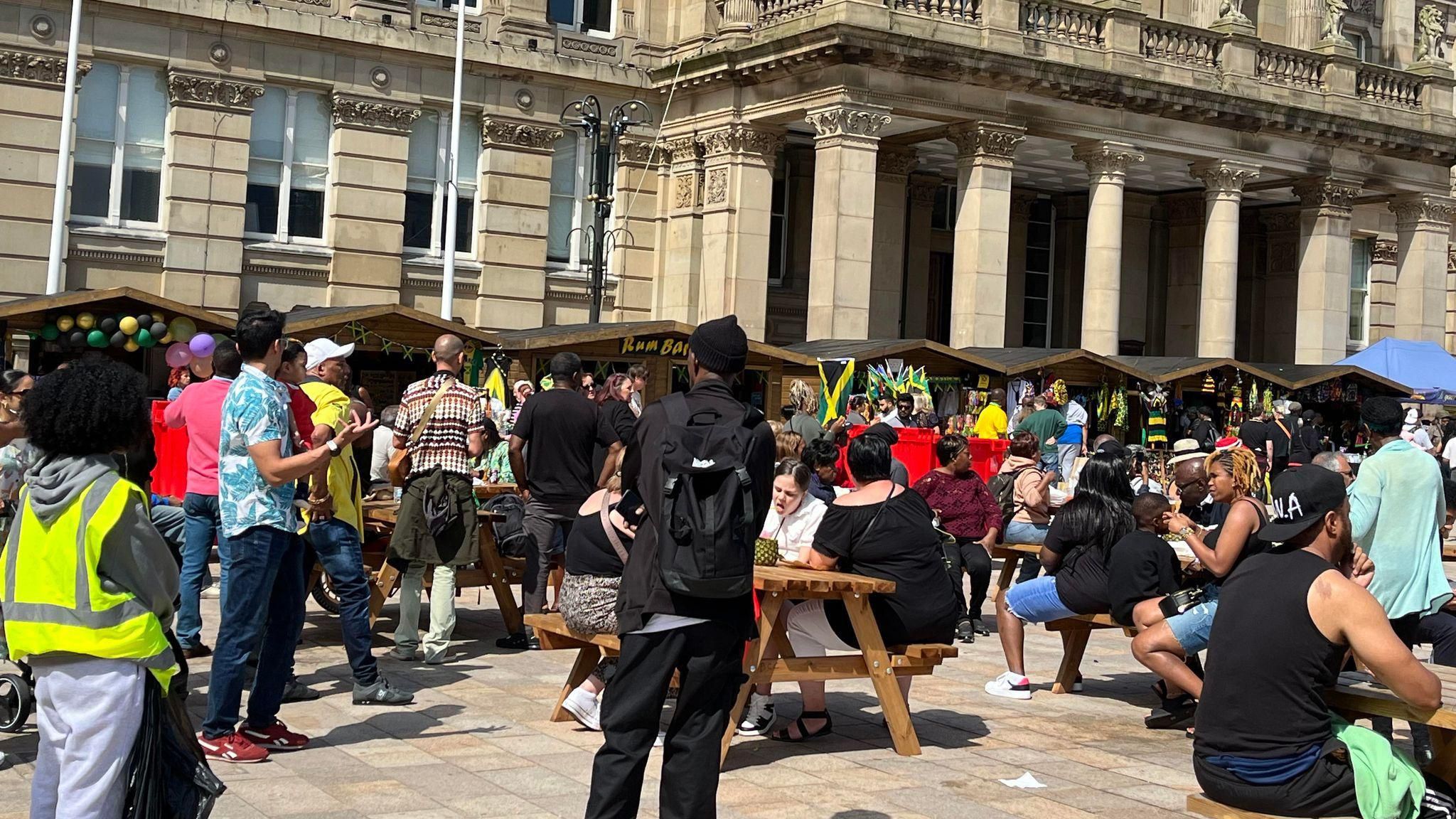 A crowd of people at the 0121 Jamaica festival, we see them thronged around park benches in front of several wooden stalls.