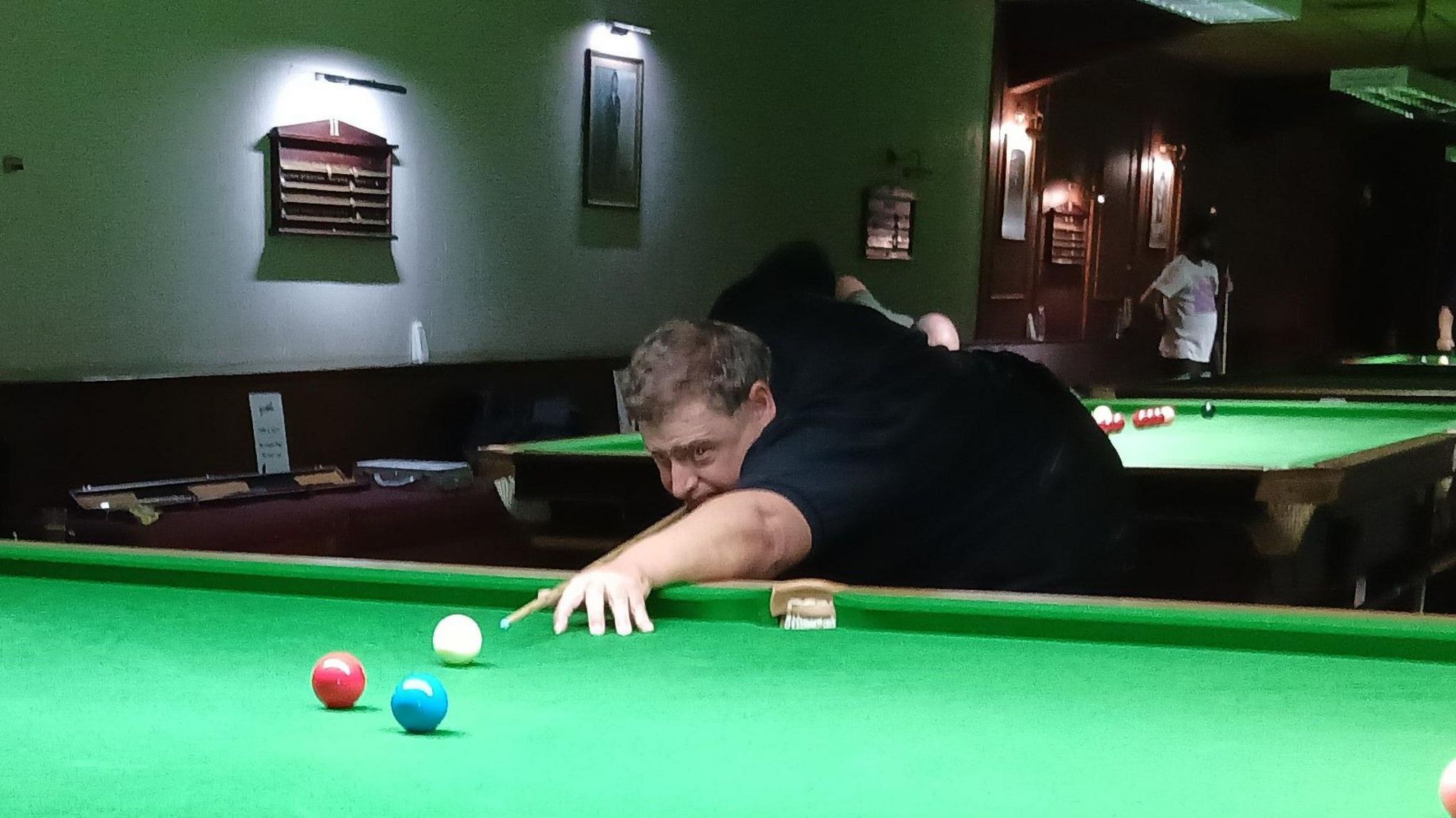 A Man dressed in black is lining up a shot on a snooker table covered in green felt. His cue is pointing at a white cue ball which is lined up to hit a red ball.