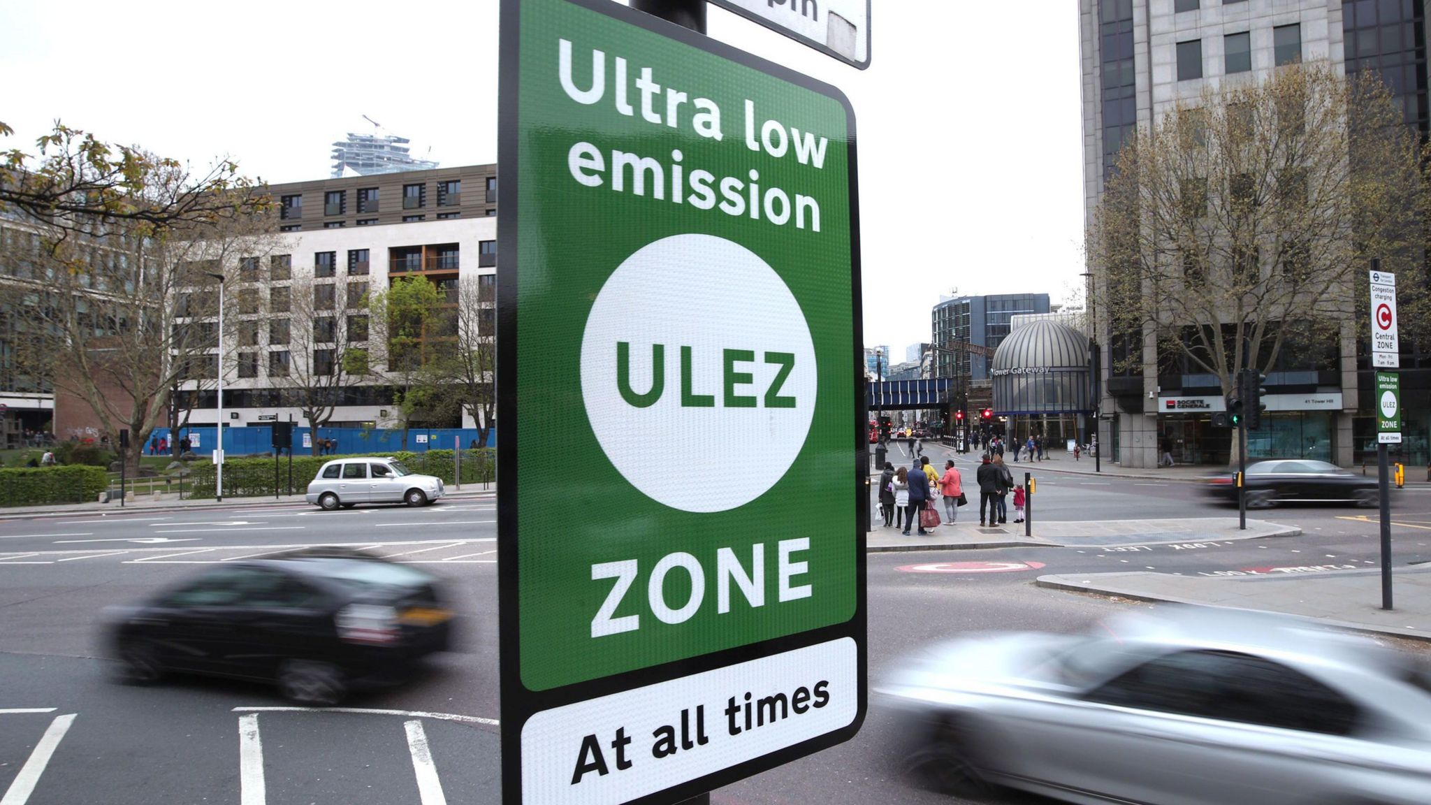 A sign, largely green, on the side of the road with ULEZ written on it and cars driving in the background