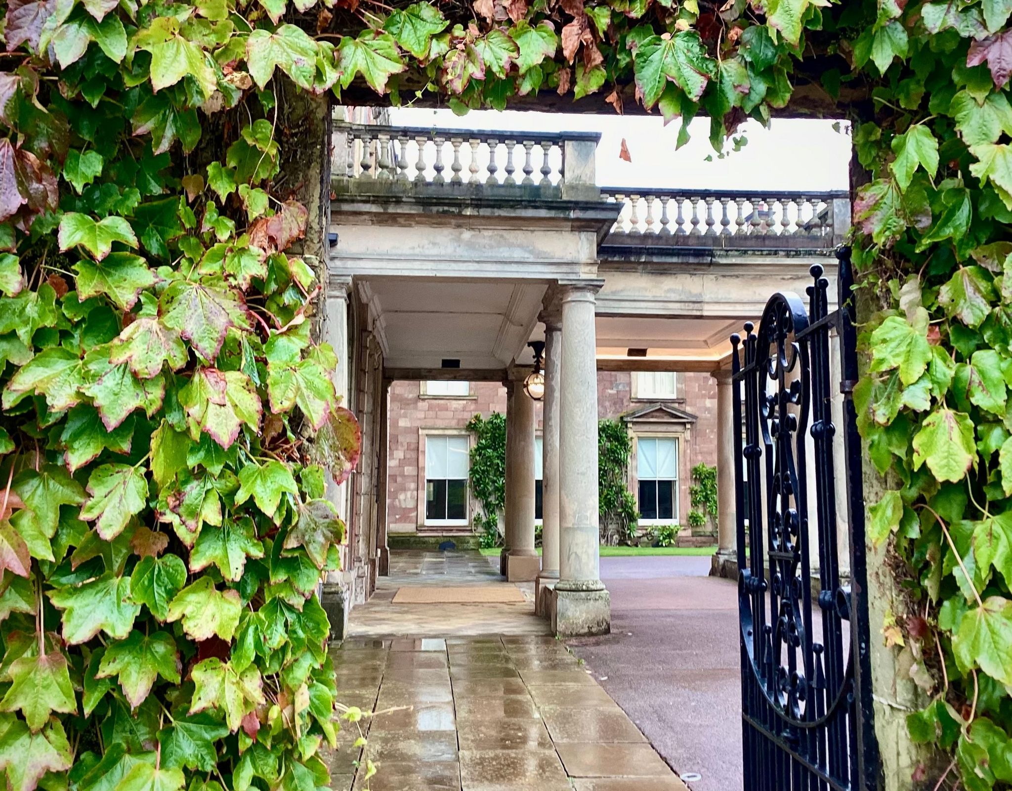 Framed by ivy, an open wrought-iron gate looks onto a pillared portico with veranda above.