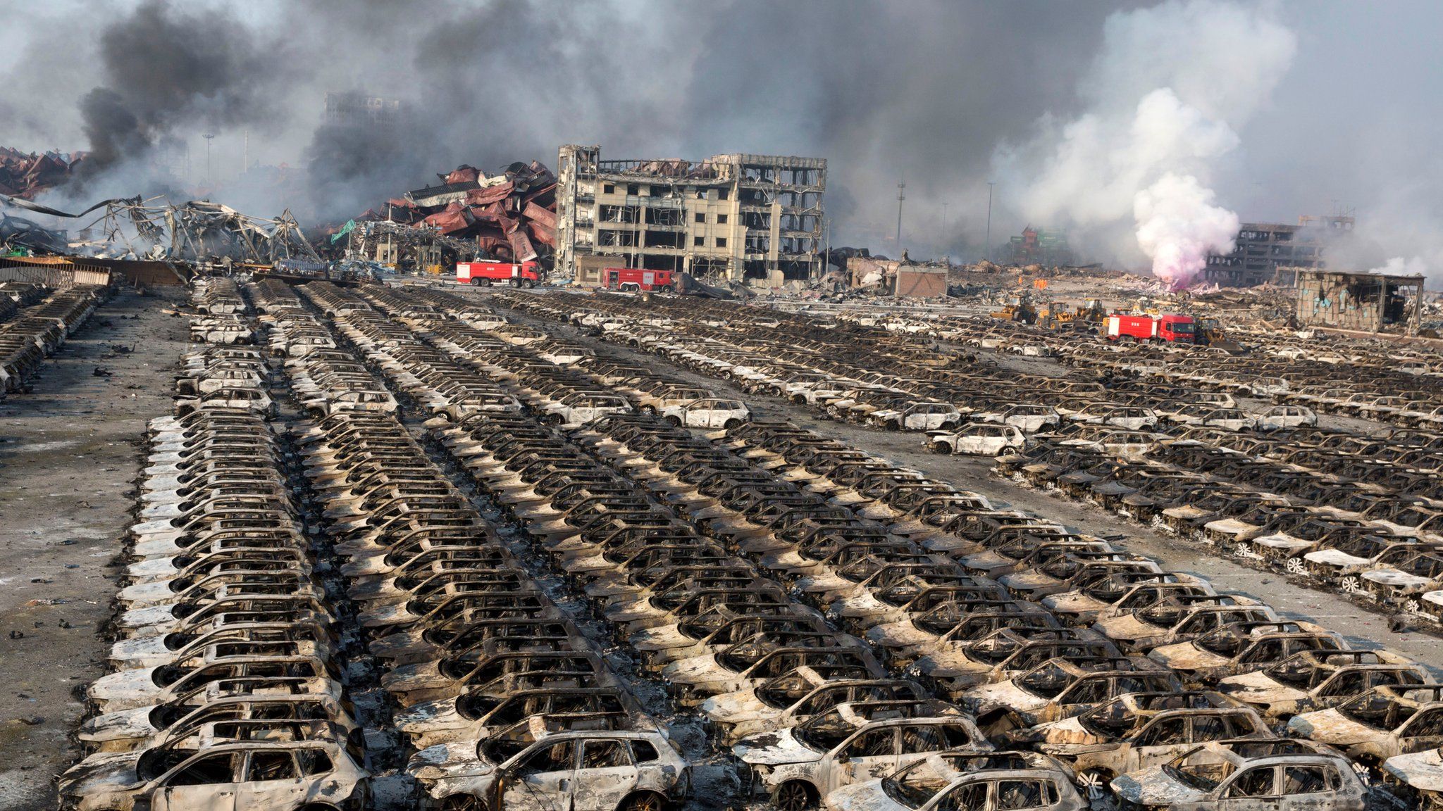 Smoke billows from the site of the Tianjin explosion, behind a parking lot filled with burnt out cars