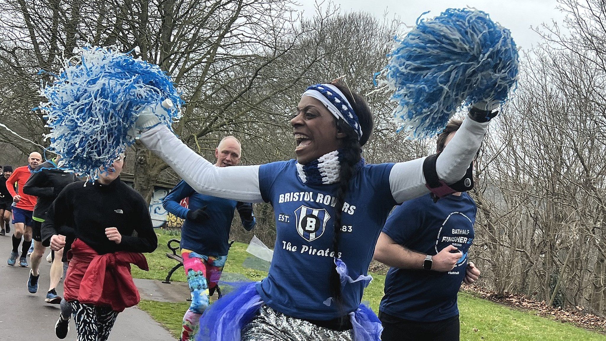 A woman wearing a Bristol Rovers t-shirt and holding pom poms runs in a park run