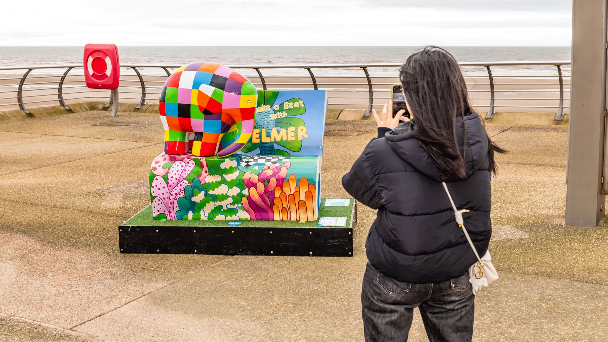 Woman takes photograph of Elmer bench sculpture on Blackpool Promenade 