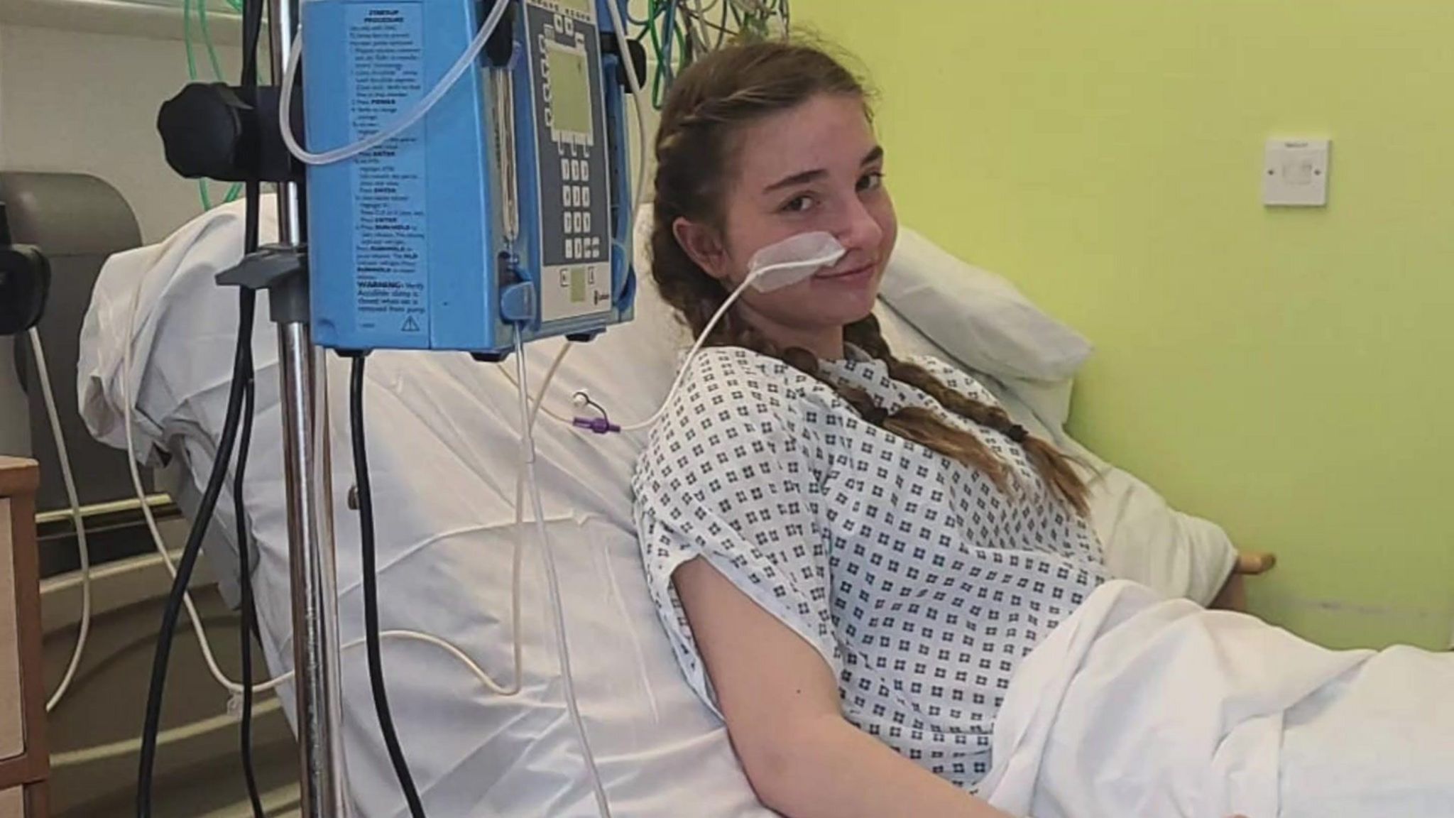 Olivia Newman-Baronius, who has long brown hair, in a hospital bed attached to a piece of medical equipment 