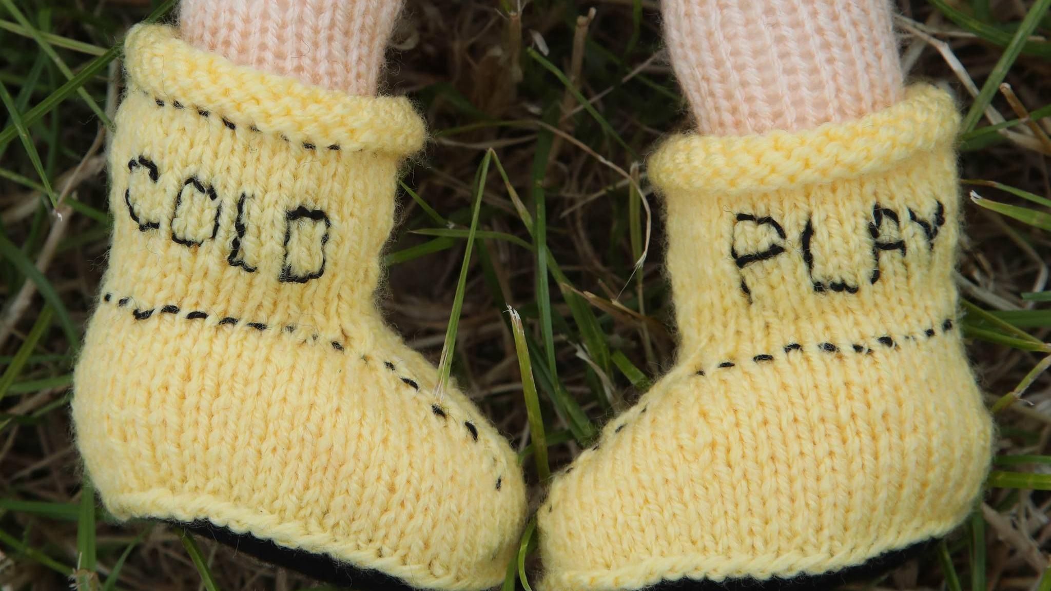 A close up of knitted yellow boots on the grass that say the words 'Cold' and 'Play' on each