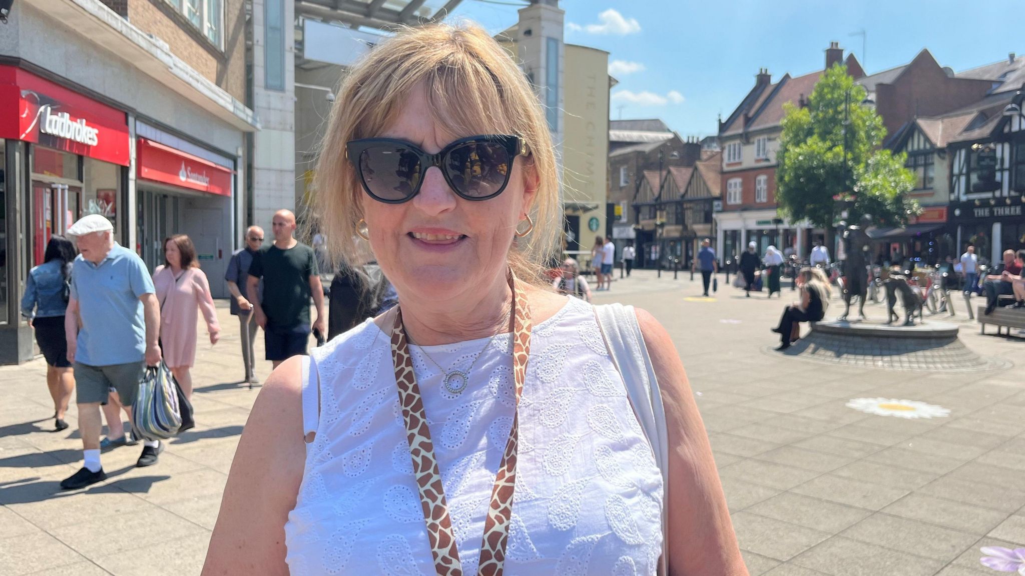 Uxbridge resident Kay who is wearing black sunglasses, a white sleeveless top and a leopard skin lanyard. In the background there is a Ladbrokes betting shop and Uxbridge town centre's paved area with a tree and some seating, as well as passers by walking past her.