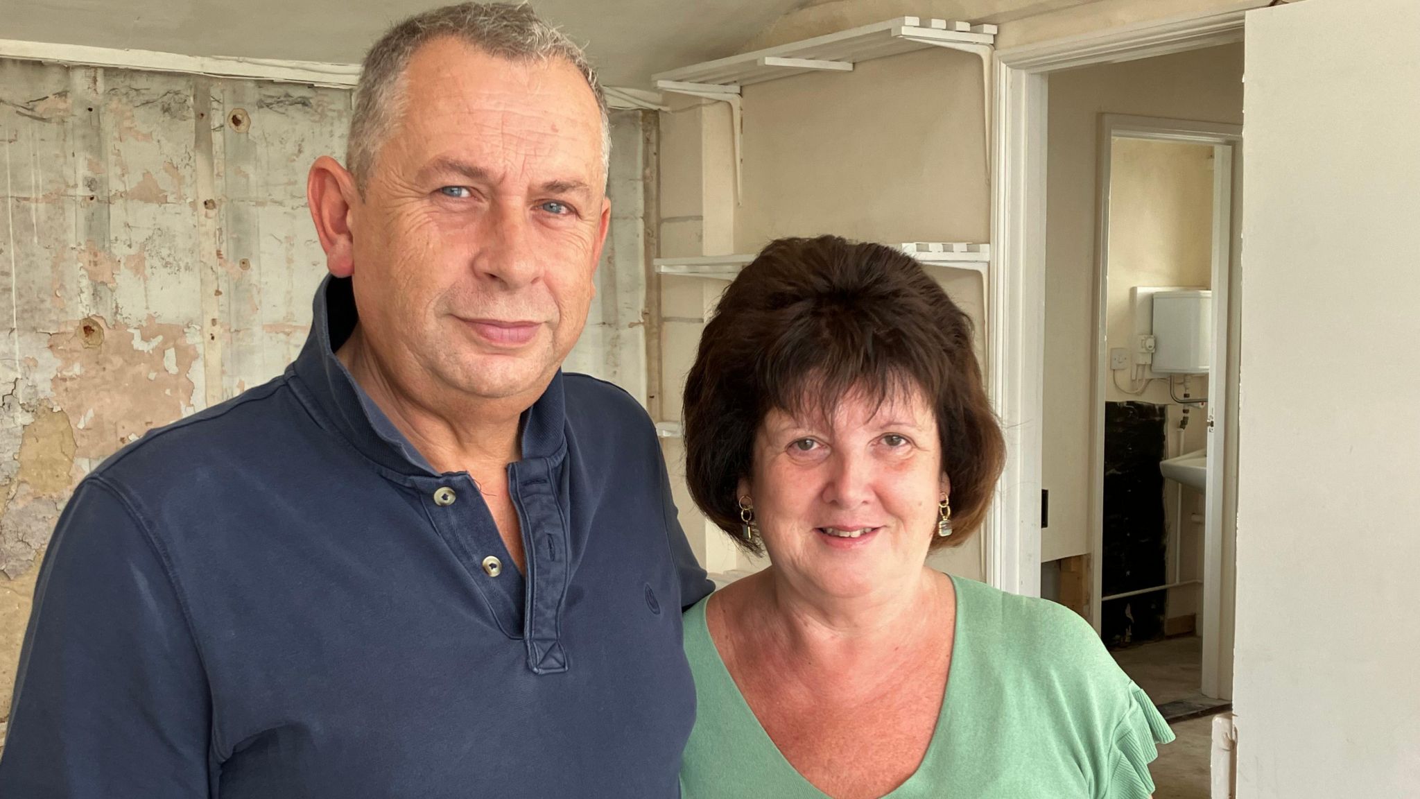 A husband and wife standing beside each other inside a shop that has been gutted following flood damage