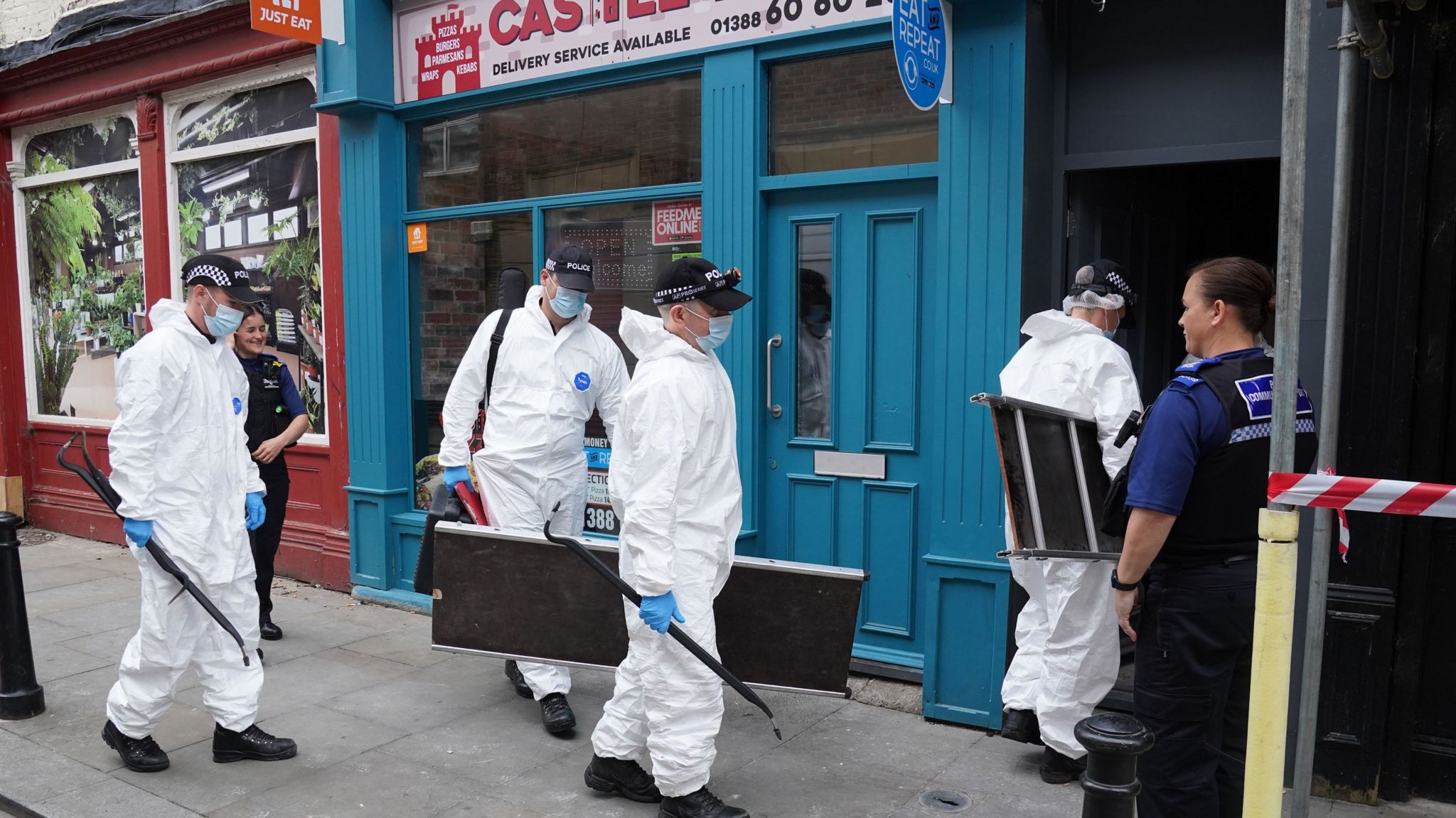 Four police forensics officers dressed in white bodysuits and wearing masks head for a doorway in a street that is guarded by a uniformed officer