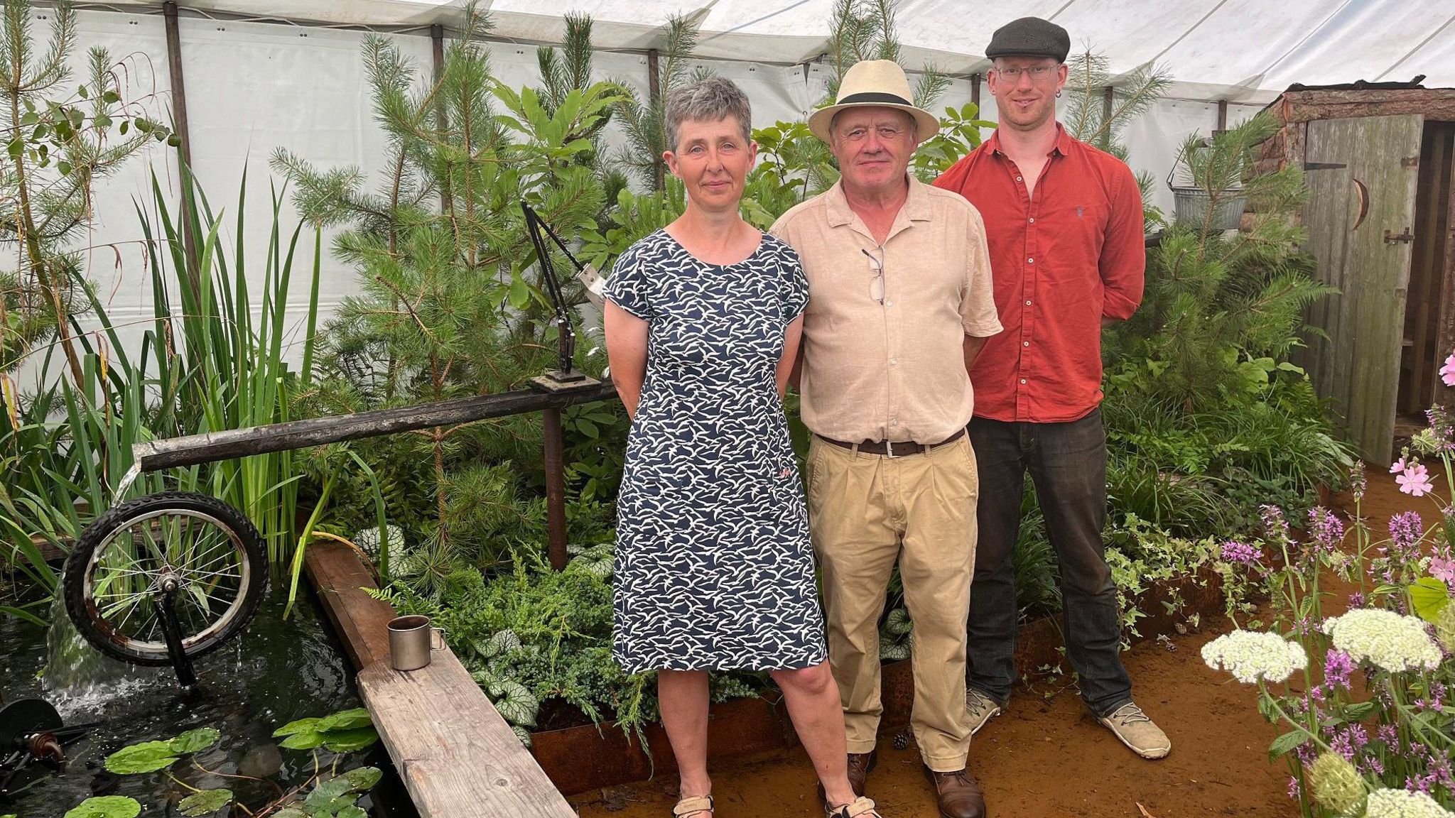 A woman and two men stand inside a show garden at Sandringham Flower Show. There's a water feature made from old pipes and a bike wheel surrounded by leafy green plants and a sprinkle of pink and white flowers in the foreground