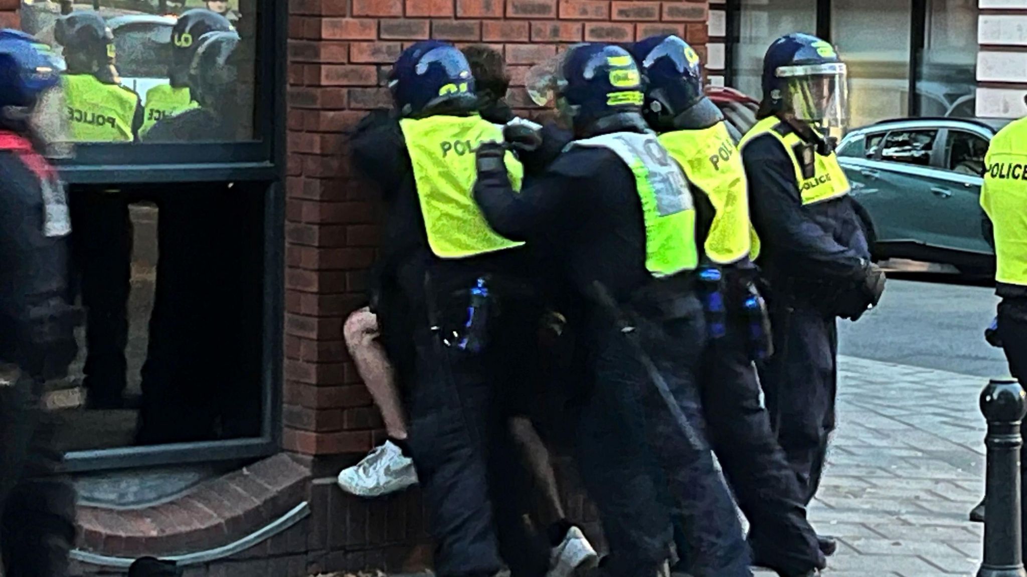 Police officers pinning a man against the wall in Bristol. The officers are wearing police uniforms and neon yellow vests and helmets. 