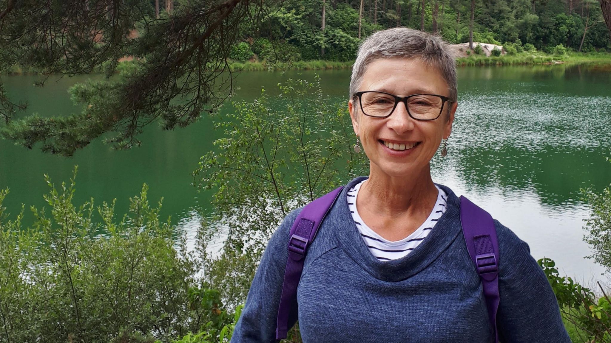 A middle-aged woman with grey hair and glasses, stood in front of a lake.