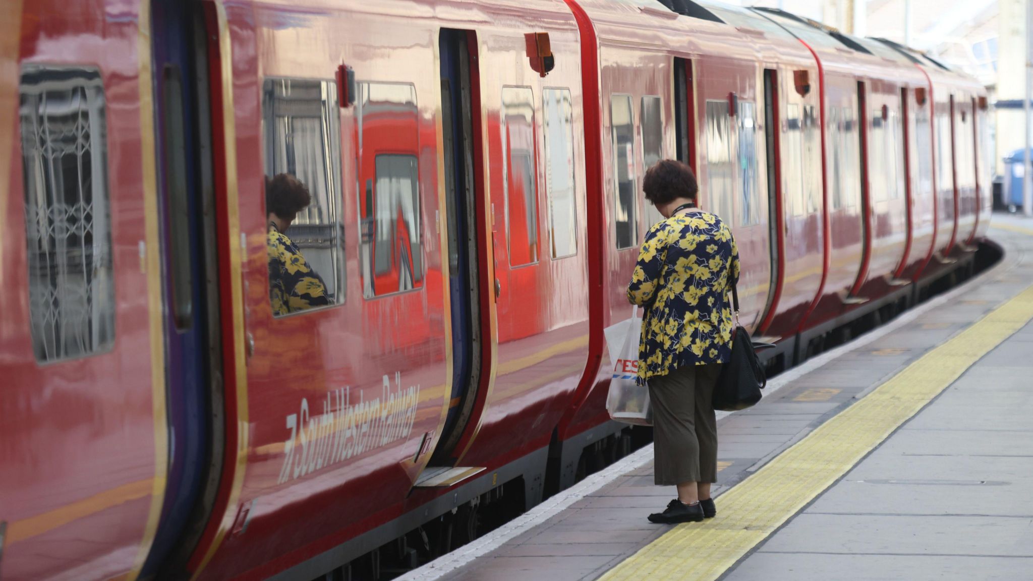 A woman stands on a platform waiting to board a South Western Railway service, in a red livery