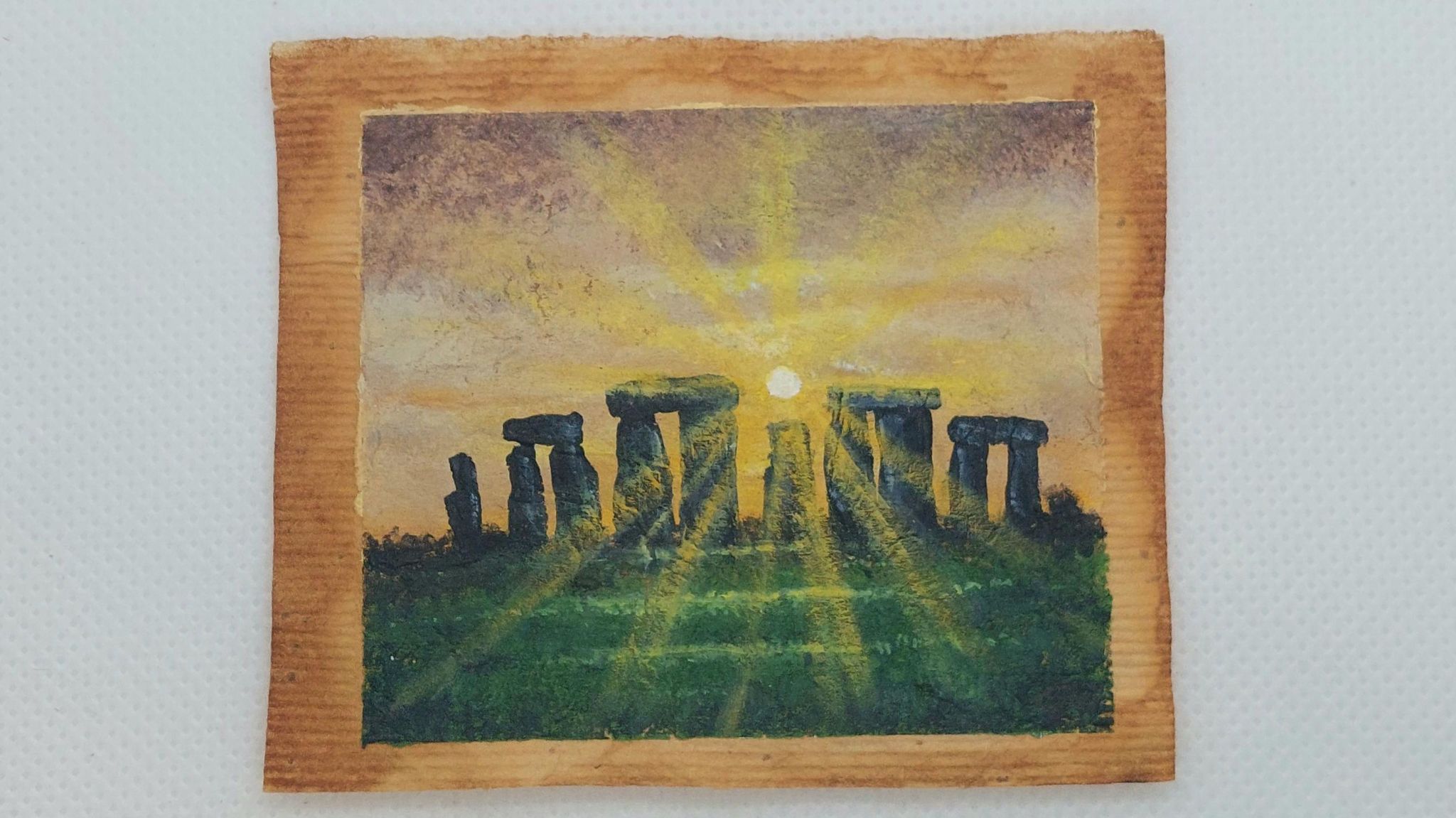 Stonehenge painted on a teabag. The sun is shining - bright yellow - through the stone circle in front which is in dark silhouette with green grass in the foreground