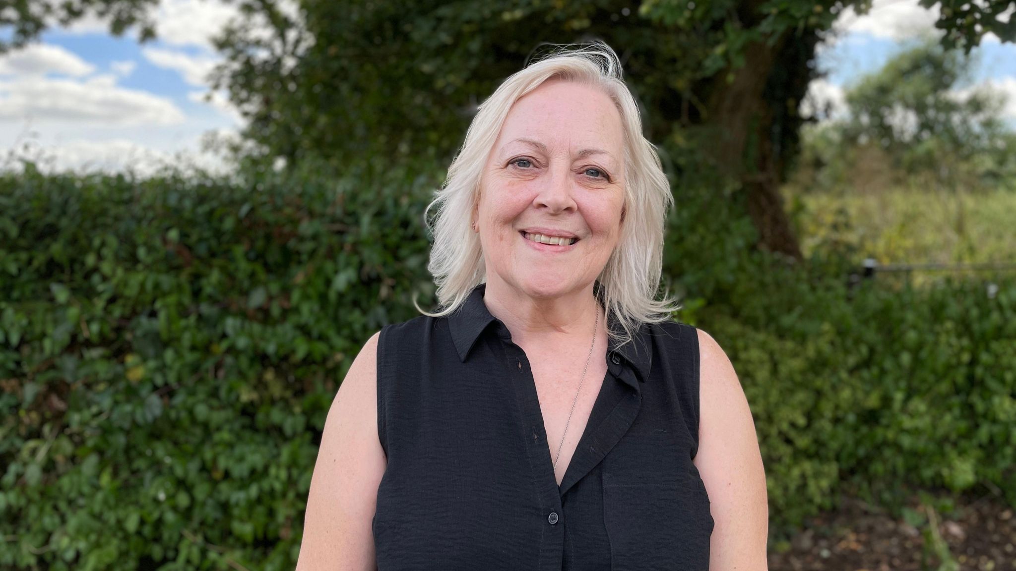 Sue Willgoss, wearing a black top, looking at the camera with a hedge and trees behind her