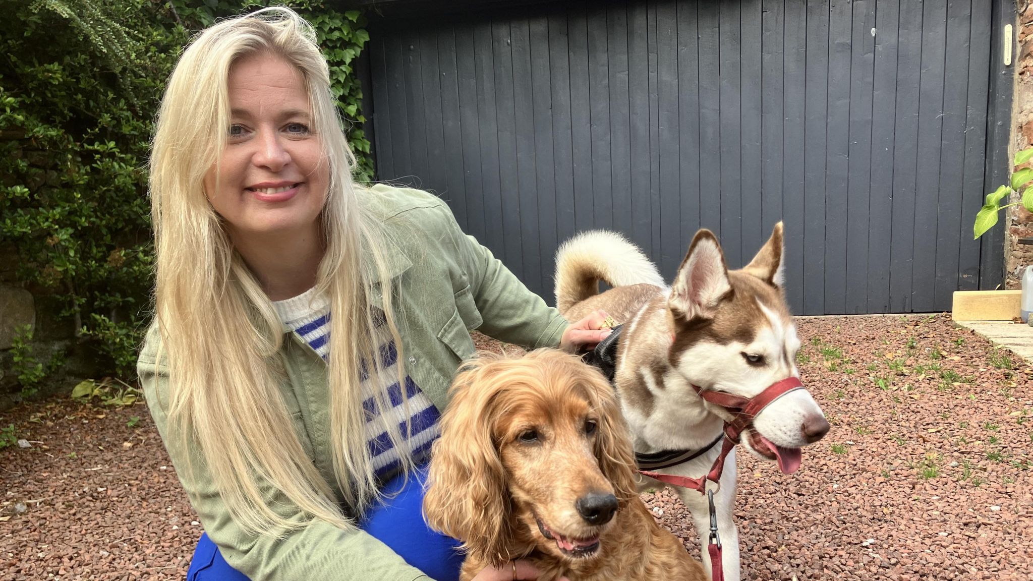 Woman with blond hair crouched down and posing with two dogs, one brown and the other white