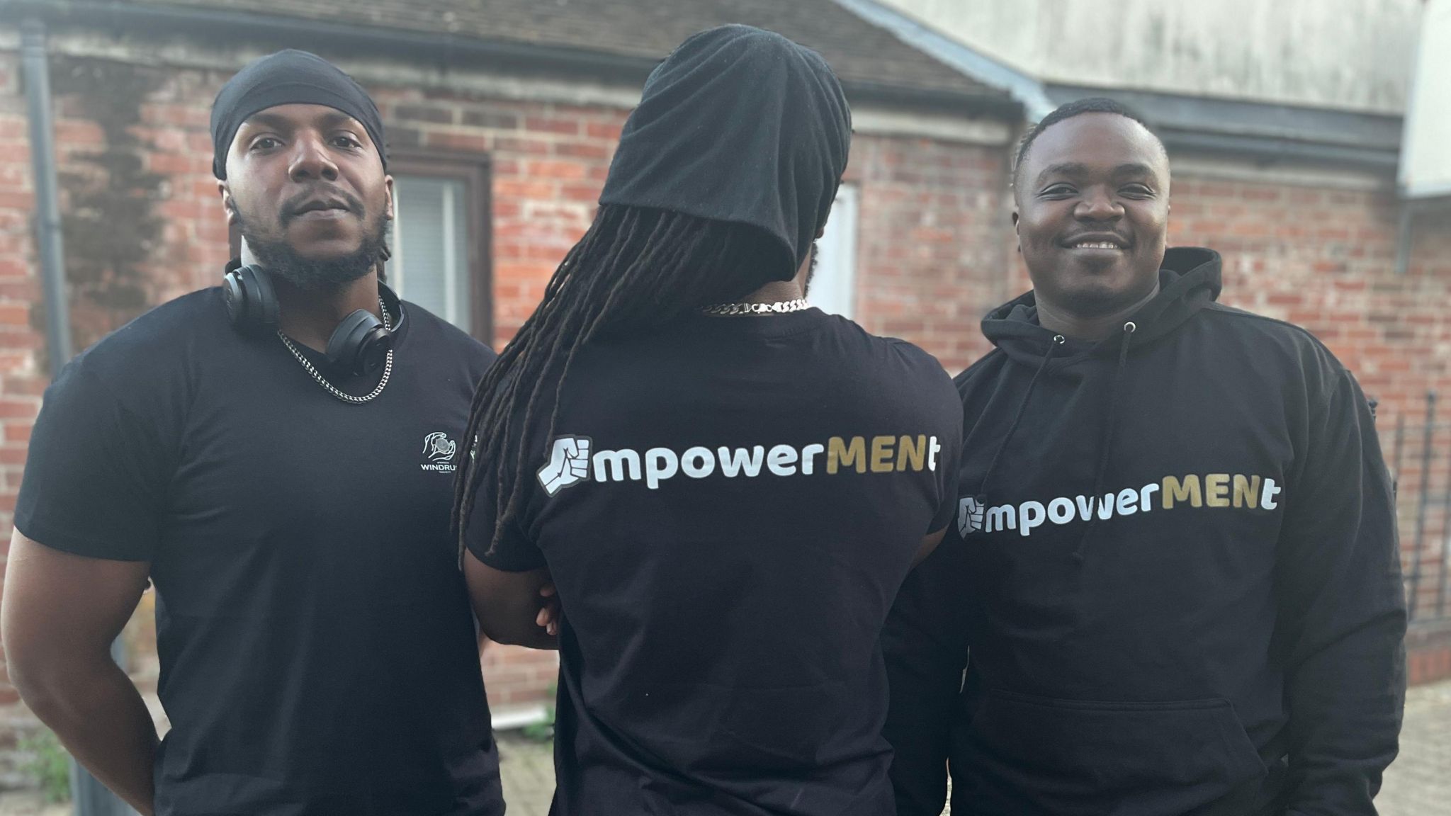 Podcast presenters known as M, K and T stand in a line, with the man in the centre with his back to the camera. They are wearing black tops emblazoned with the EmpowerMENt logo