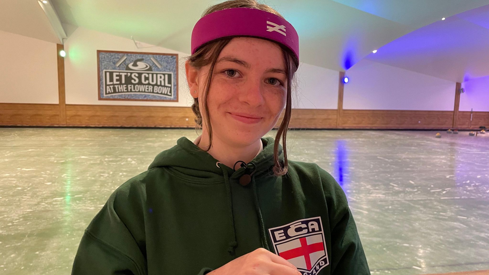 Marinna smiles at the camera, wearing a cerise head band and a green England Curling Association hoodie, with the ECA logo featuring the English flag. The ice rink, with a sign saying 'Let's curl at the Flower Bowl' and an illustration of a curling weight and two brushes, can be seen behind her.