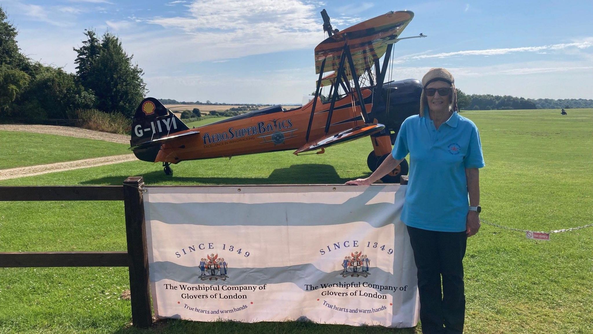 Catherine Grimley looking at the camera with the RFC Rendcomb field and a biplane behind her. Mrs Grimley is wearing a brown hat, blue t shirt and black trousers.