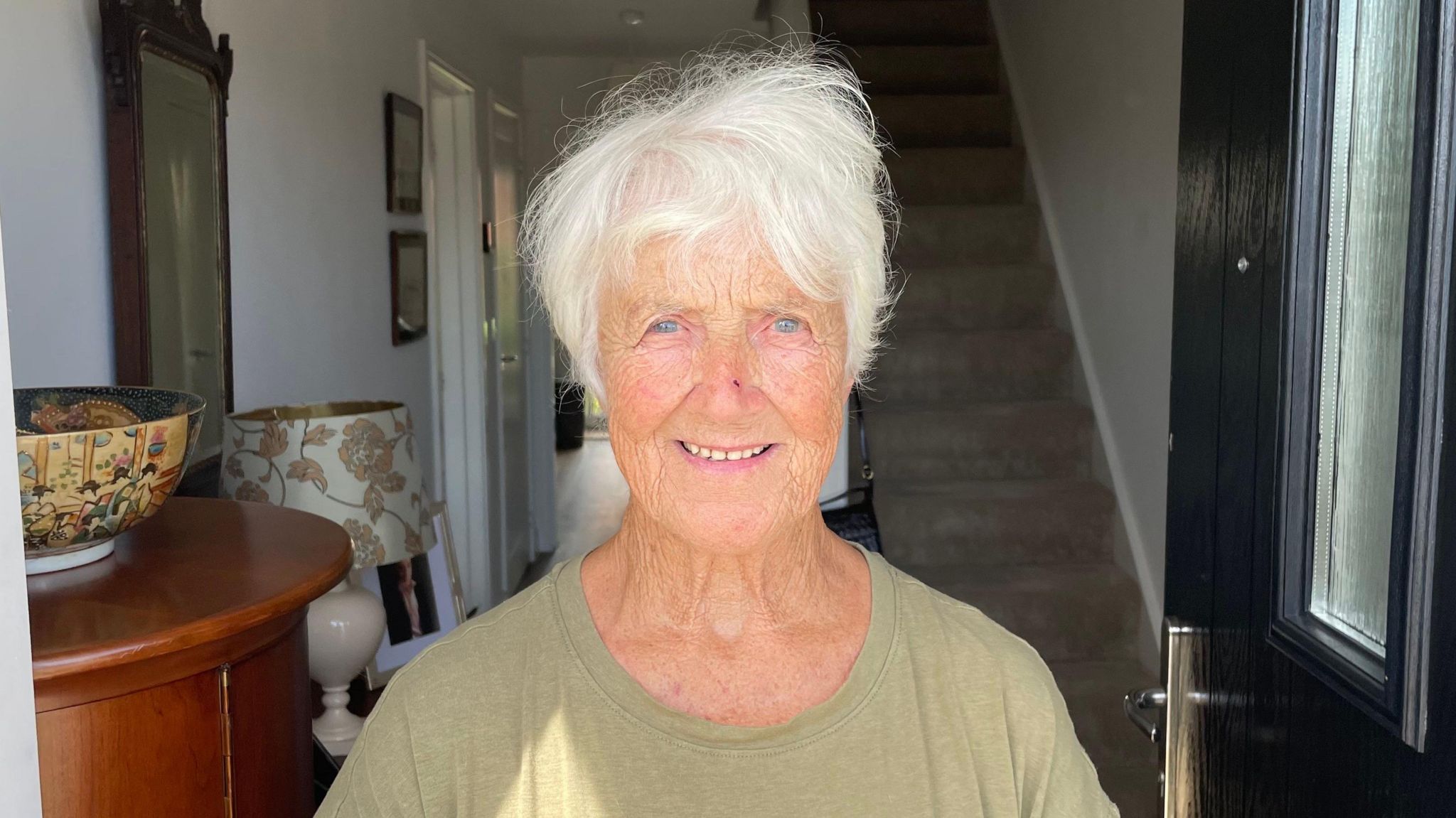 Looking direct to camera, Josephine Jackson, with white hair and blue eyes, is standing in the doorway of her new home in Bacton. She is smiling and wearing an olive green loose-fitting top.