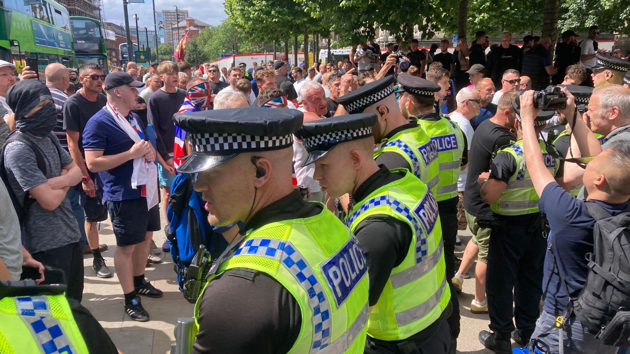 A line of police officers, wearing hats and reflective police vests stand between two opposing protest groups. One protester has the flag of Great Britain draped around his shoulders, while another protester is wearing a scarf around his face with a hat on