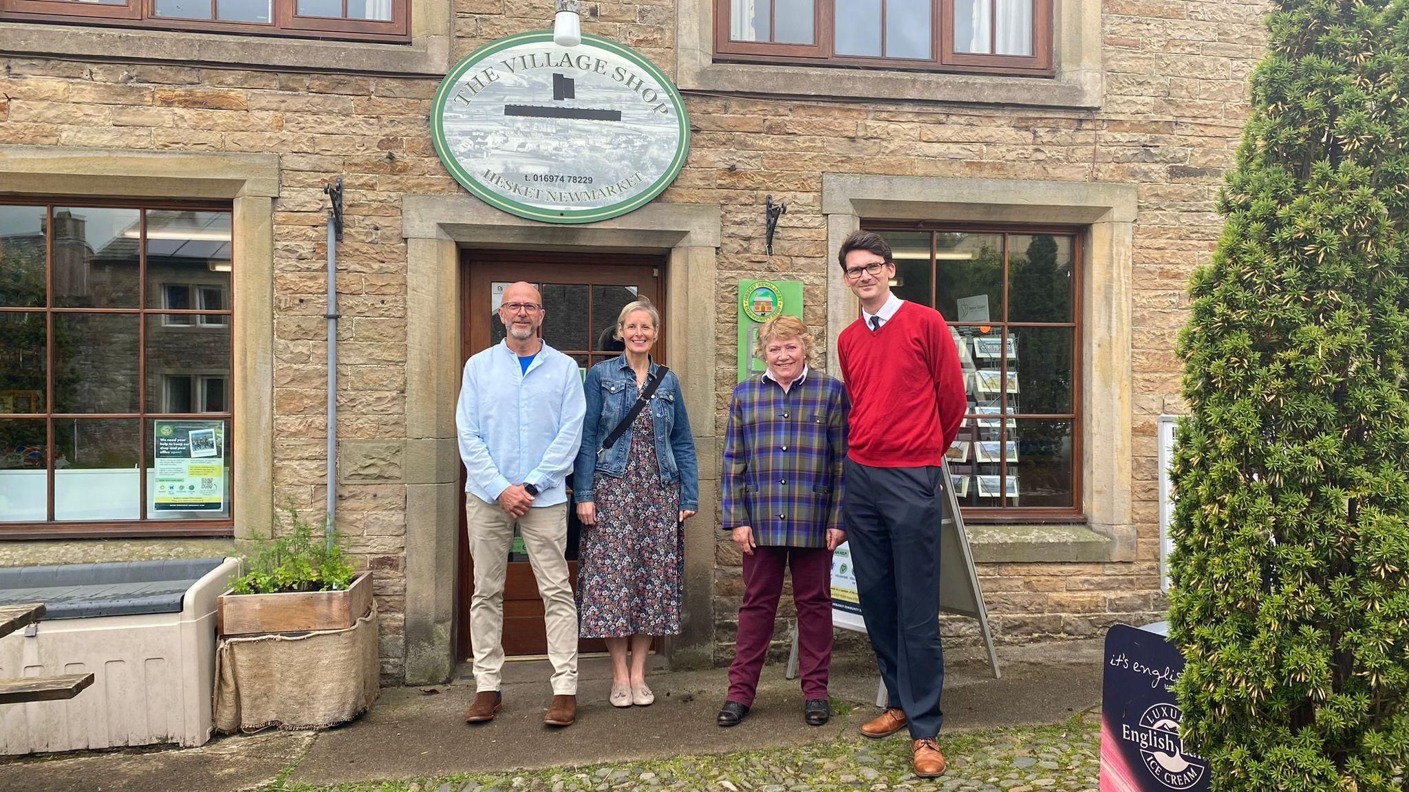 Three members of the Hesket Newmarket Community Shop Project with local MP Markus Campbell-Savours in front of the old brick village shop. Left to right, a man with a bald head and short, neat grey beard and glasses, wearing a light blue, buttoned-up shirt over a dark blue t-shirt, light tan trousers and dark tan shoes; a woman with short, light hair wearing a blue denim jacket over a long flowing dress and with a bag strap showing diagonally across her front; a second woman with short, blonde hair wearing claret trousers and a buttoned shirt in a purple, yellow, green and blue tartan pattern; and a very tall man with short dark hair and glasses, wearing a bright red jumper over a white collared shirt and dark tie.