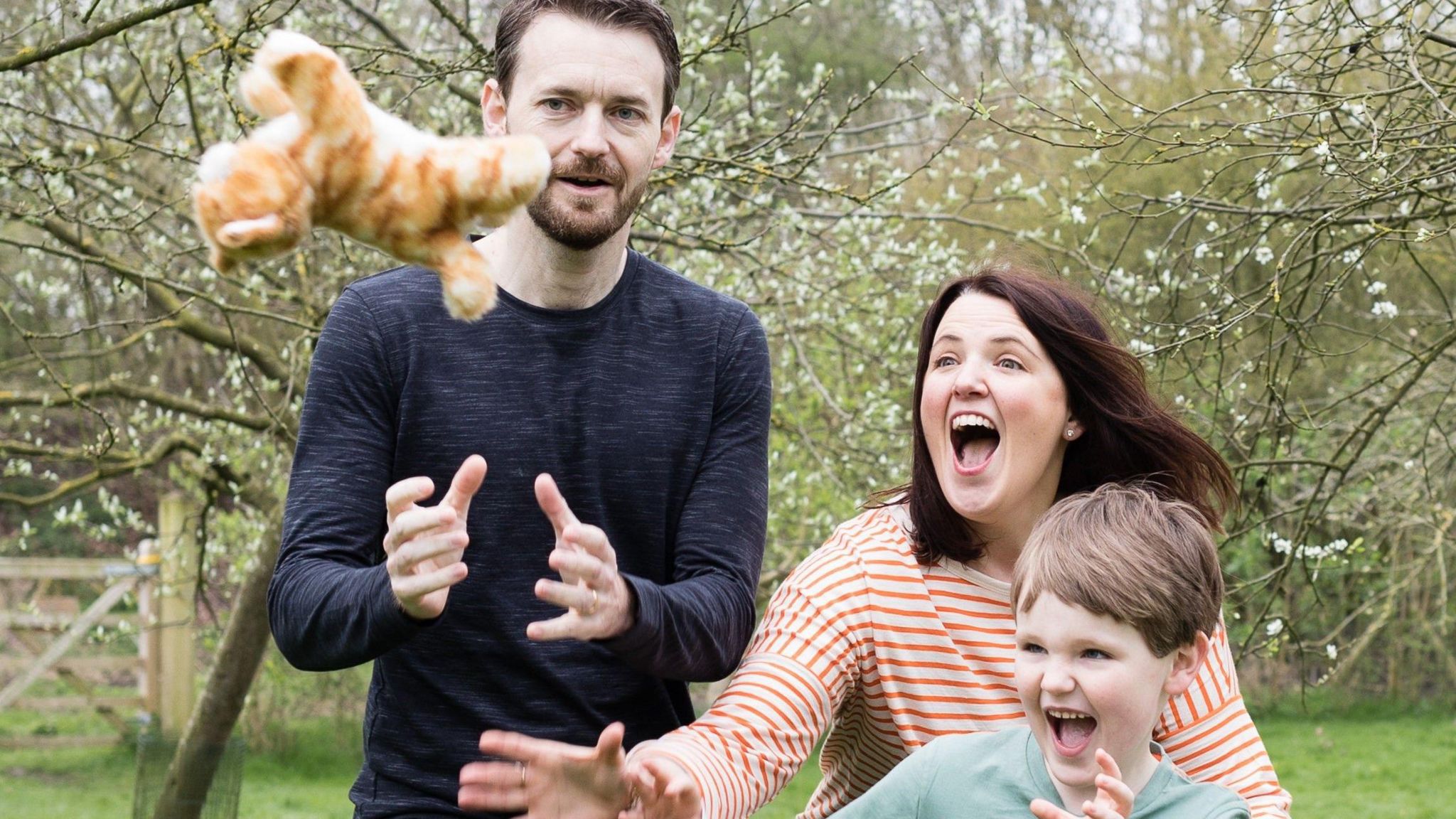 Cliff Gibson in a dark blue jumper with Rachel Gibson in an orange stripped top and and their son aged about eight or nine in a light blue top, all trying to catch an orange stuffed toy, standing in a field with blossom trees behind them