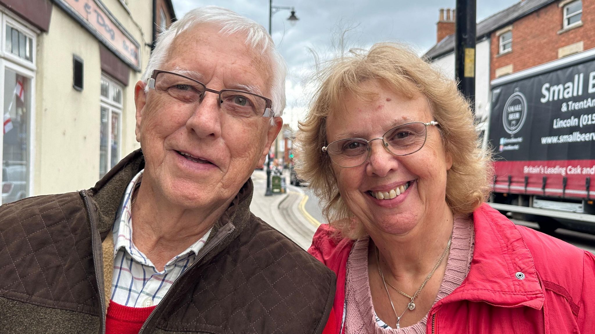 Brian Price on the left with wife Sandra. Both are smiling for the camera and Brian is wearing a brown jacket and has grey hair, while Sandra is wearing a pink coat and jumper. They are both stood in the street