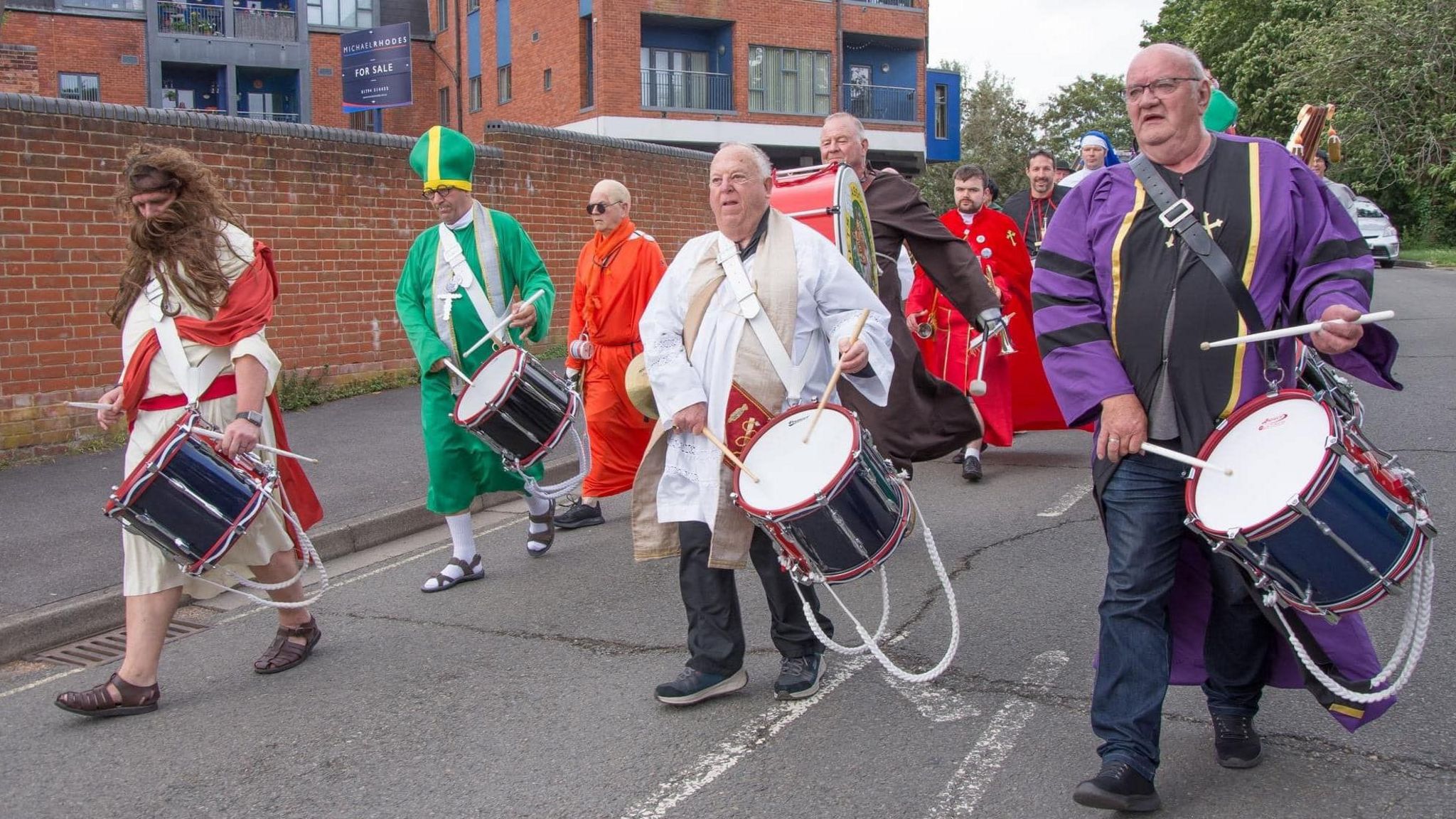 Several men dressed in different fancy dress costumes walk along the road carrying drums.