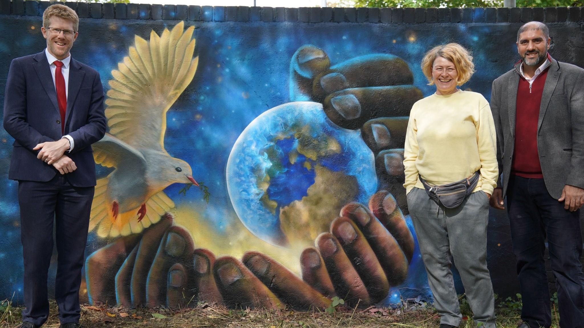 Two men and a woman stand to the sides of a mural depicting several hands cupping planet Earth and a dove of peace against a dark blue background punctuated by stars.