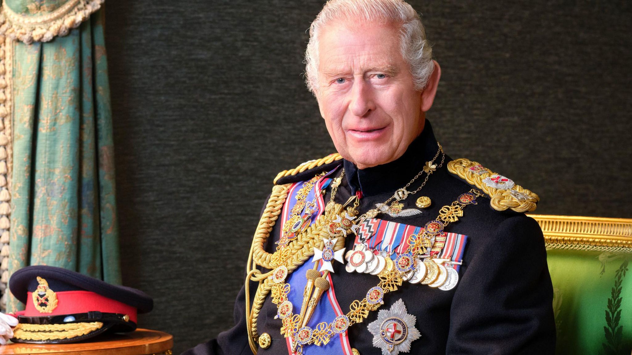 King Charles III sitting in the Grand Corridor at Windsor Castle, wearing his Field Marshal No1 Full Ceremonial Frock Coat with medals, sword and decorations. His Field Marshal cap, white gloves and baton are placed on the table beside him.