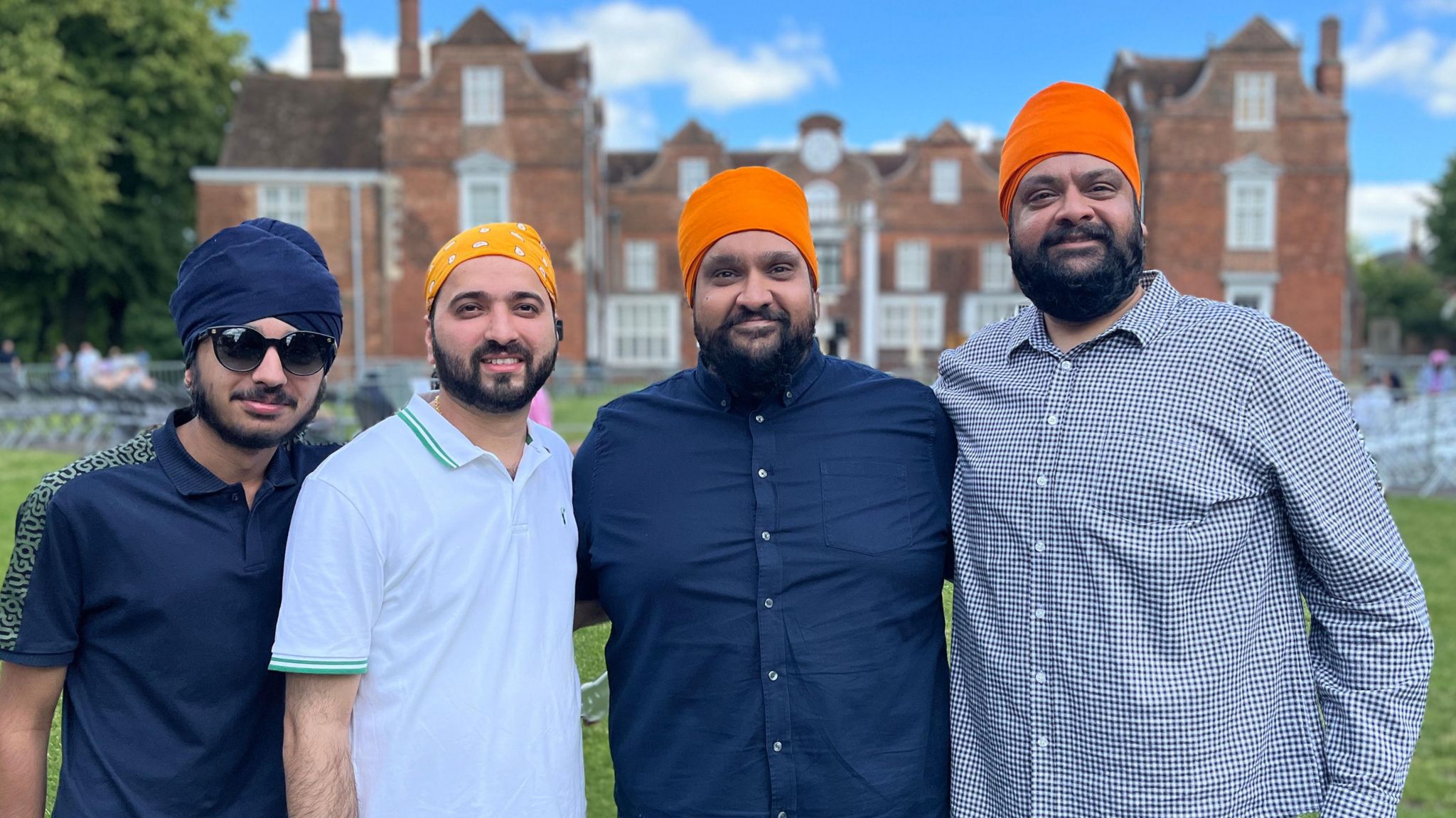 Four Sikh men, wearing blue, yellow and orange turbans, stand in front of a Tudor mansion