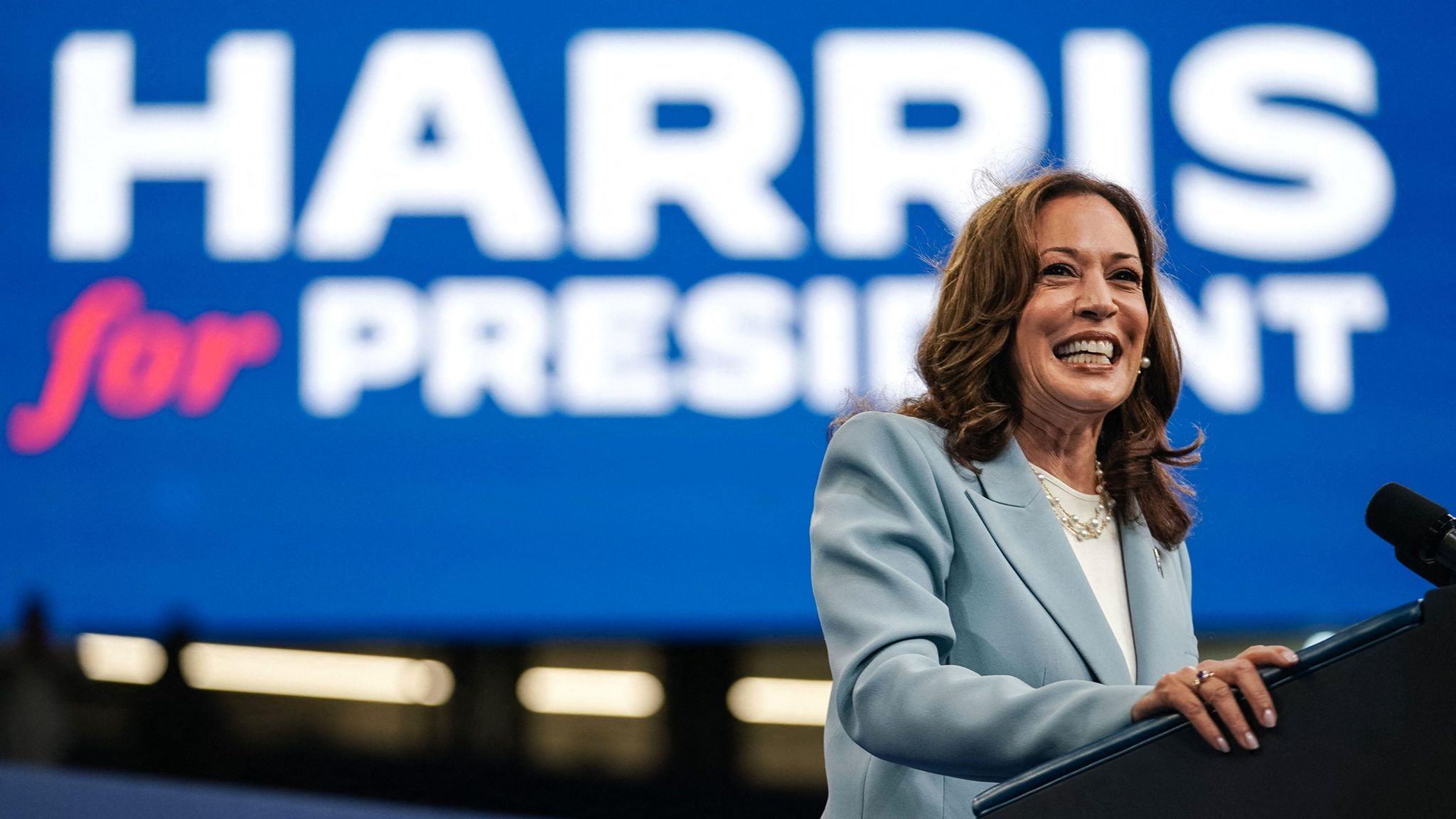 Kamala Harris in a blue suit jacket standing at a podium in front of a poster that reads "Harris for president"