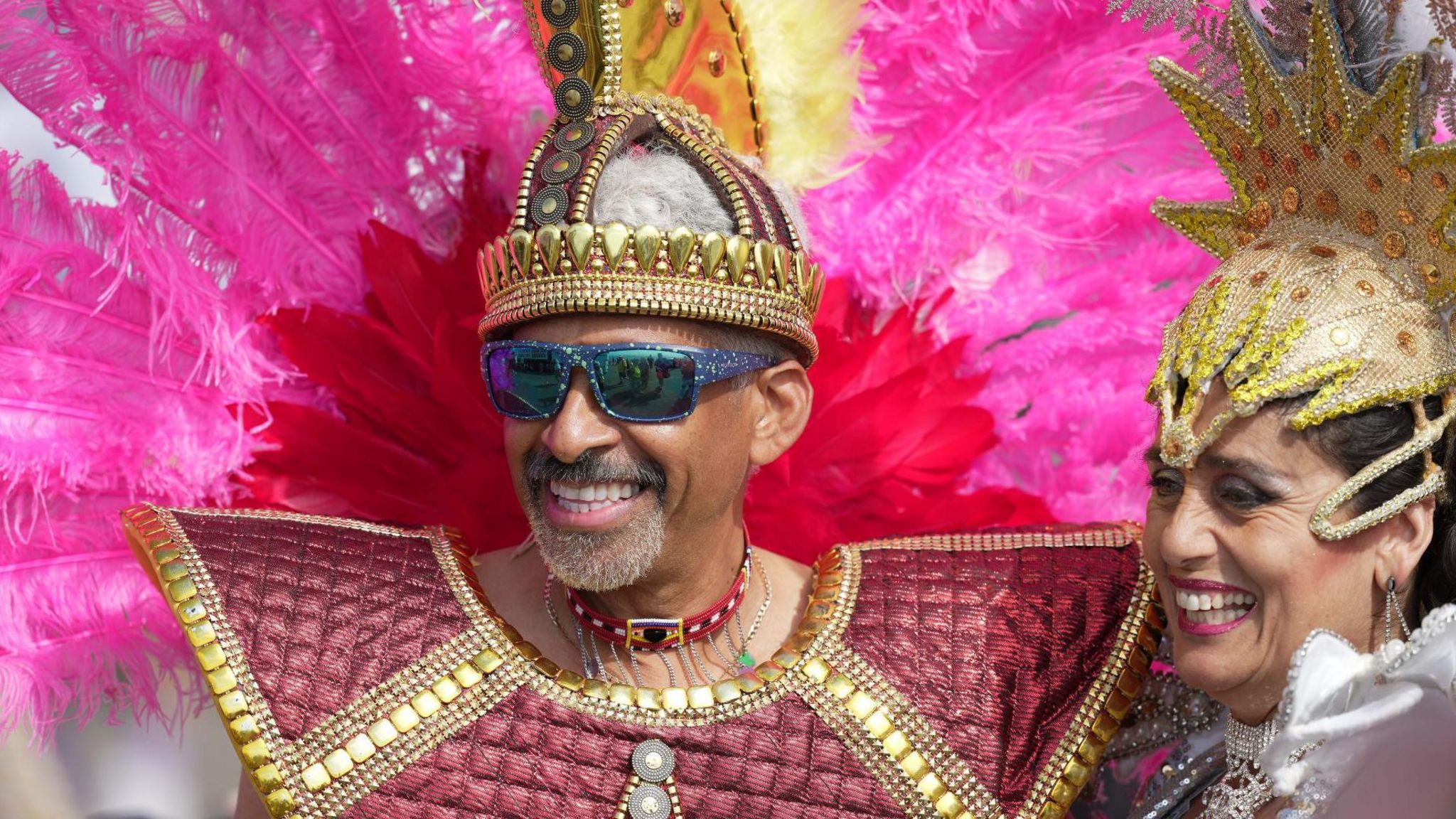 Two people wearing golden headdresses. Man on the left, woman on the right. The man on the left hair a large pink feather tail behind him