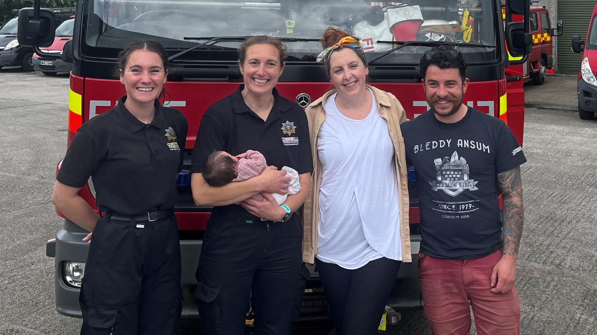 Two women firefighters stand in front of a fire engine, with one holding baby Olive, standing next to Olive's parents. All are smiling 