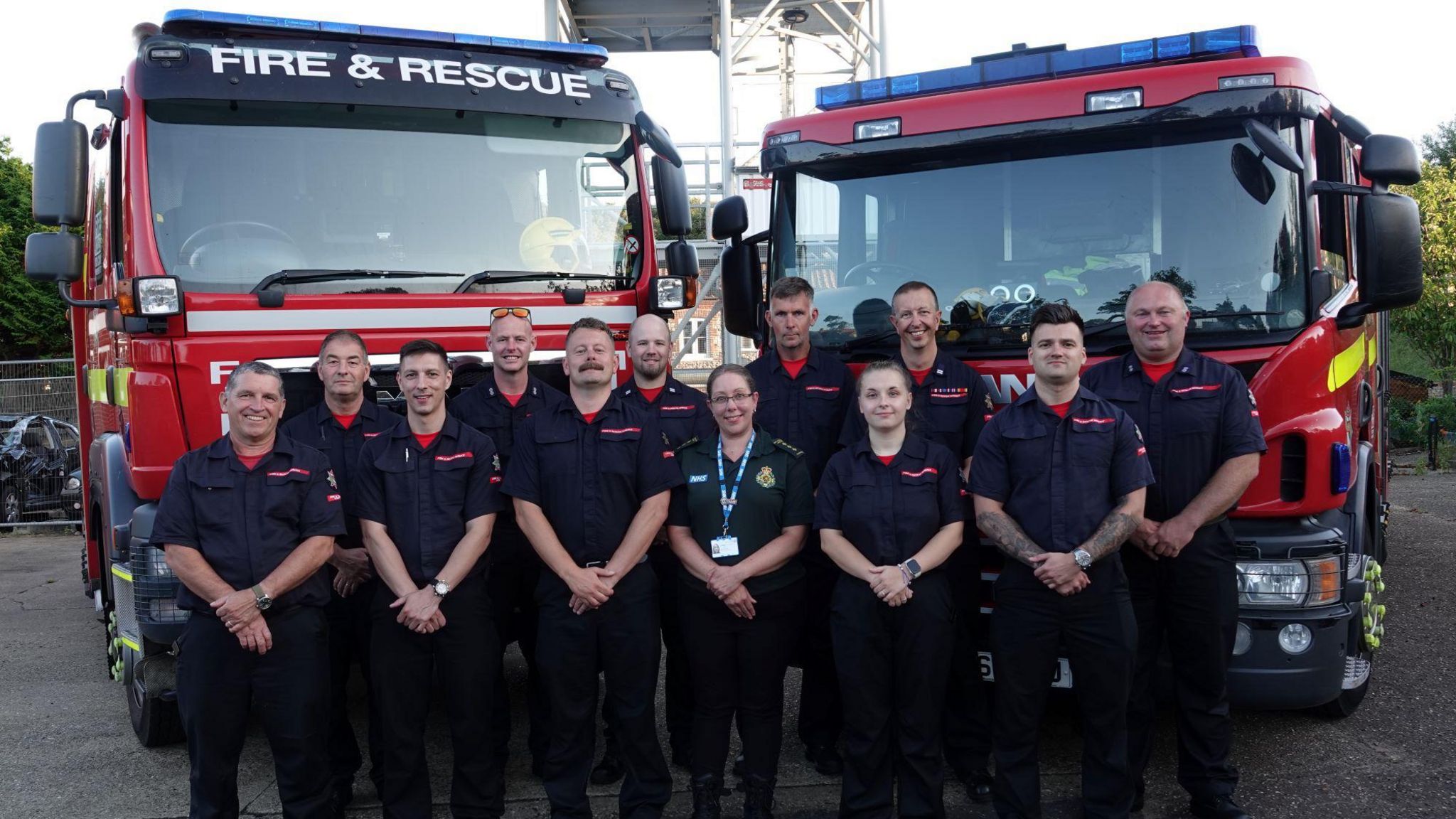 Firefighters in uniform stand with Vikki Darby from EEAST in front of two red fire engines. 