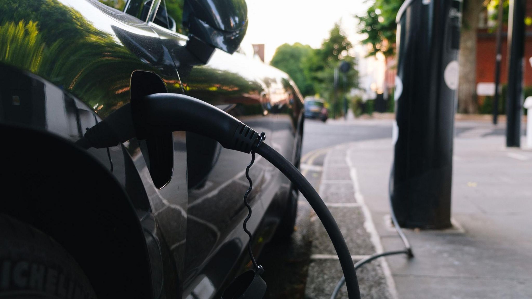 A black car parked next to a pavement with an electric charger plugged in. It is connected to a black post by a wire.