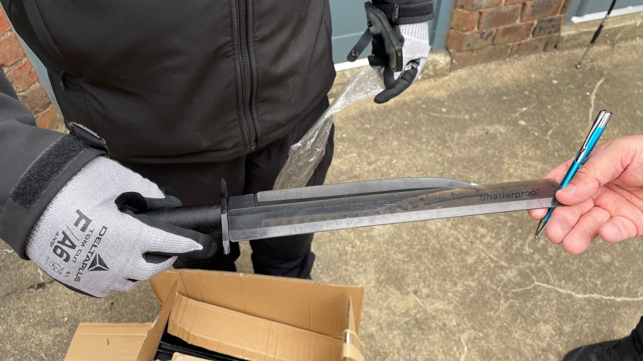 A police officer holds a zombie knife in a gloved hand, while another officer holds a 30cm rule against its blade. The officer with the ruler is also holding a pen. Part of a cardboard box can be seen on the concrete floor, while some plastic wrapping can be seen held in another gloved hand.