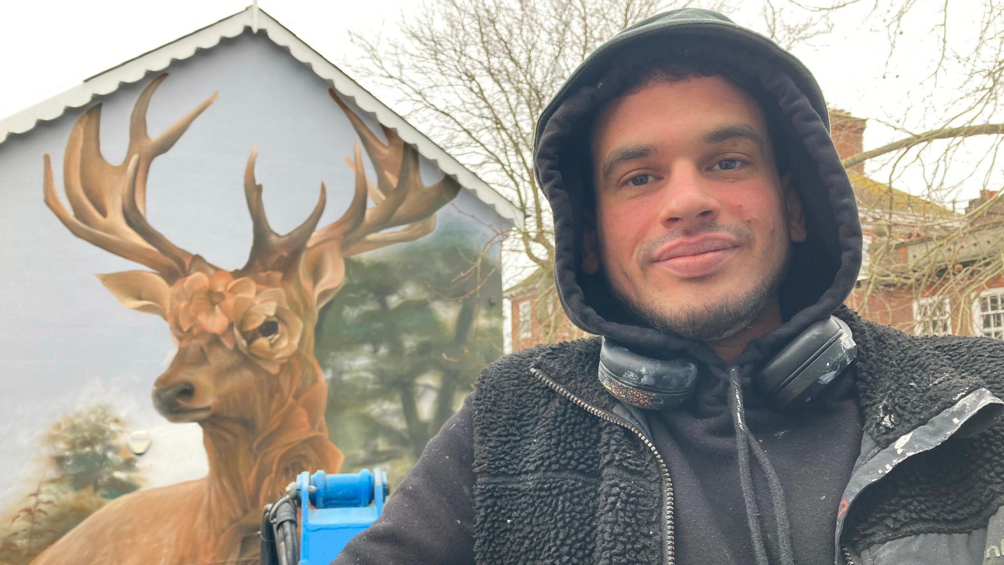 A man wearing a hooded jumper with headphones around his neck stood in front of a wall with a deer painted on it.