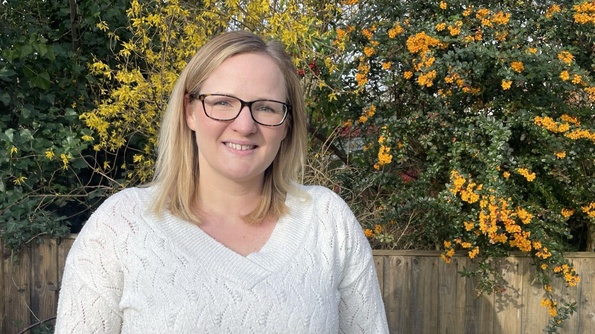 A woman wearing glasses and a white jumper stood in front of a wooden fence.