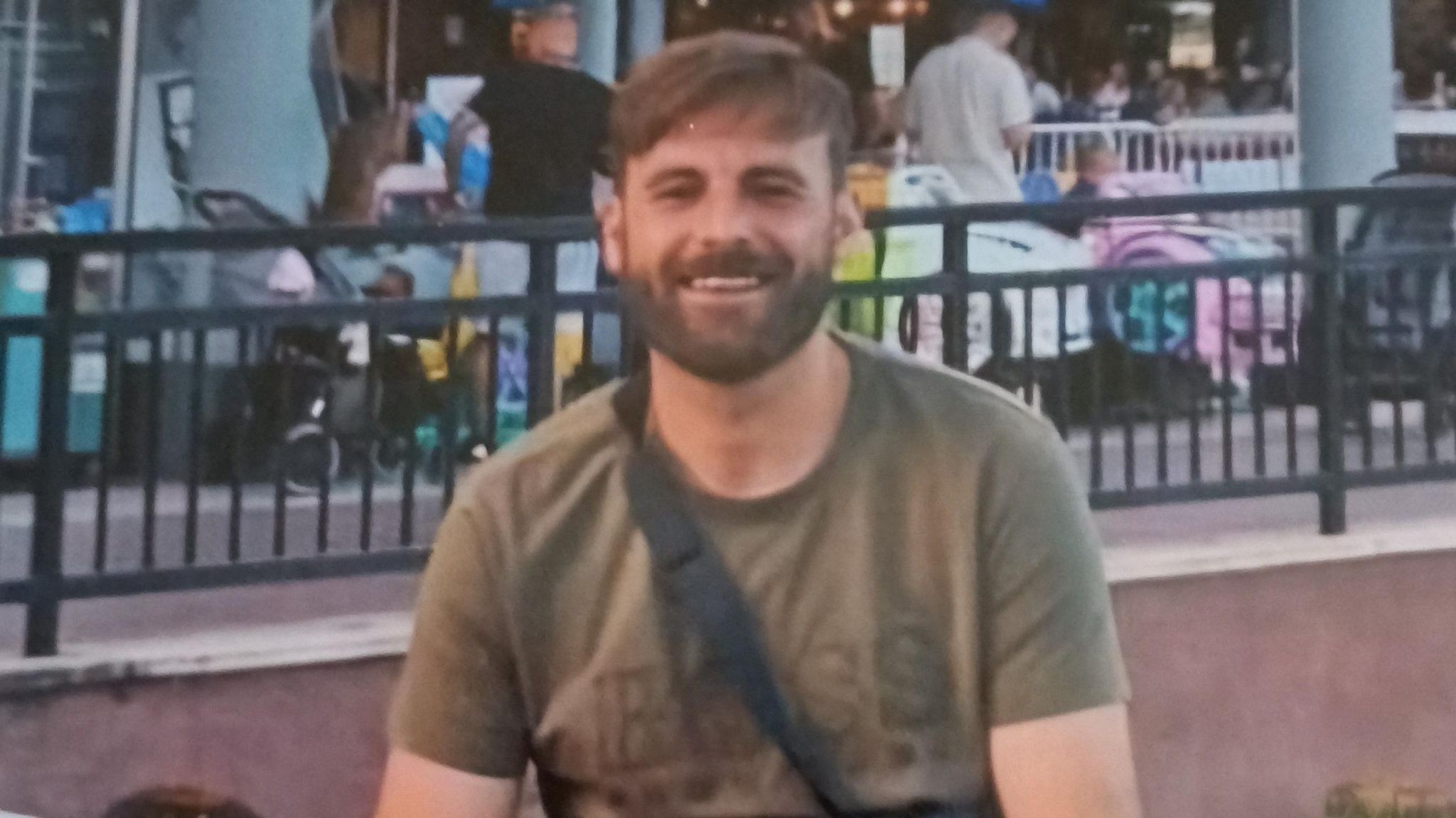 Christopher Jones wearing a green t-shirt. He has brown hair and a beard and is sitting outside a row of restaurants