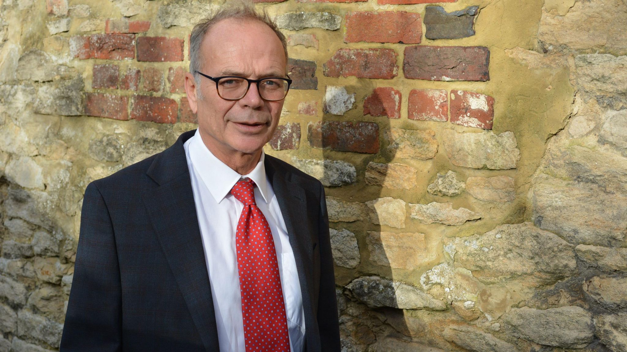 Dr Simon Opher standing next to a wall. He is wearing a white shirt, red tie and black jacket