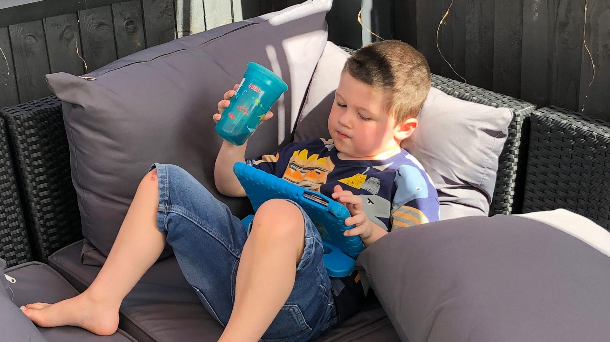 Toby is pictured lying on a garden sofa while holding a cup and watching a show on a tablet device